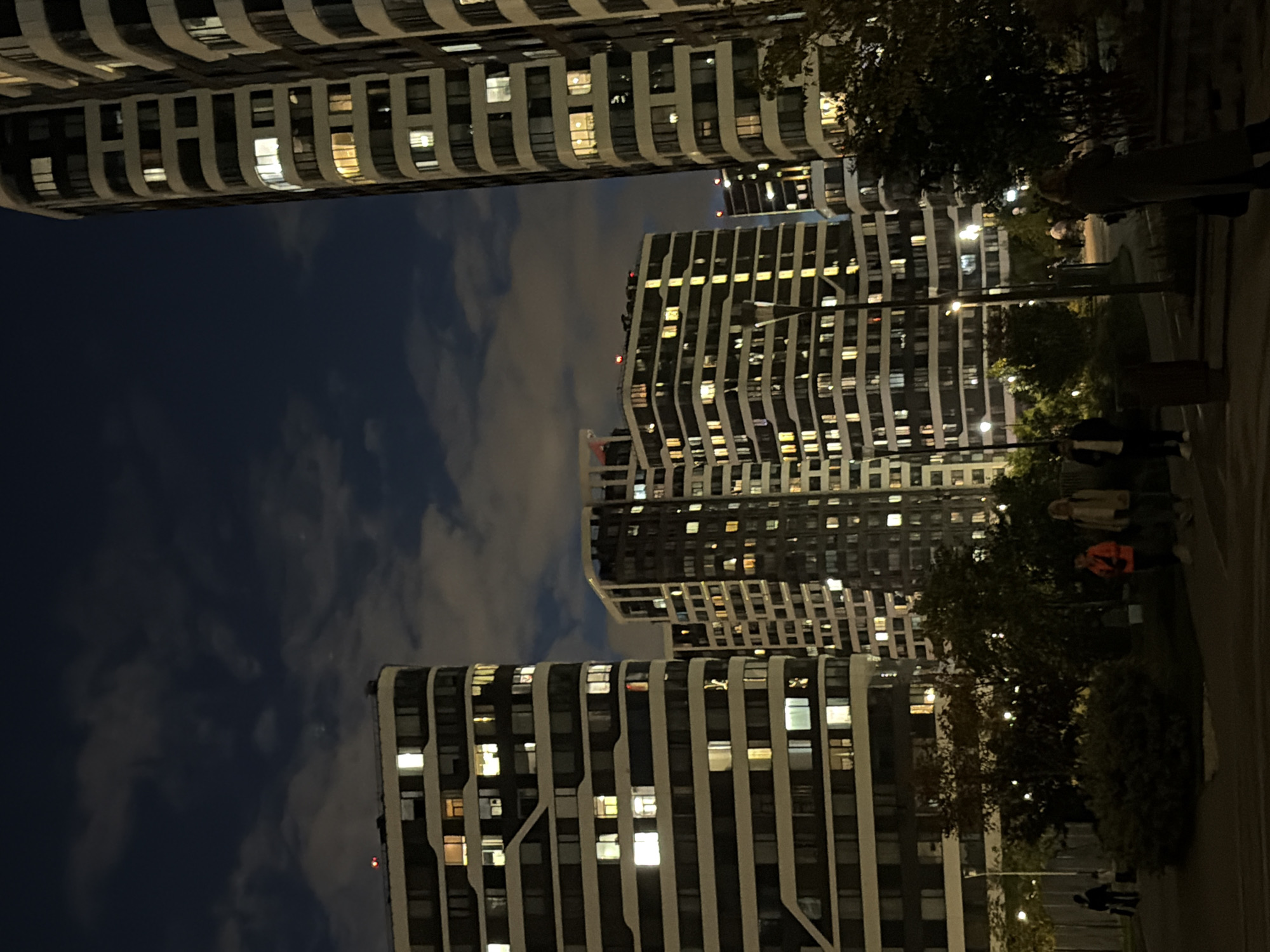 The image depicts a night scene in a modern urban residential area in Moscow, Russia. The foreground features a paved walkway with some people visible. Two individuals are standing and looking away from the camera, one wearing an orange jacket and the other a beige jacket. Further back, several other figures are seen walking or standing. A trash bin is present on the left.
The midground is occupied by a park-like setting with trees and lampposts. The trees are illuminated by faint lights, suggesting they are part of the urban lighting scheme.
The background is dominated by several tall, multi-story apartment buildings with a contemporary architectural design. The buildings are lit from within, with many windows glowing with warm light, indicating residential occupancy. The sky above is dark with some clouds, consistent with nighttime. The overall impression is one of a well-lit, modern residential complex at night.