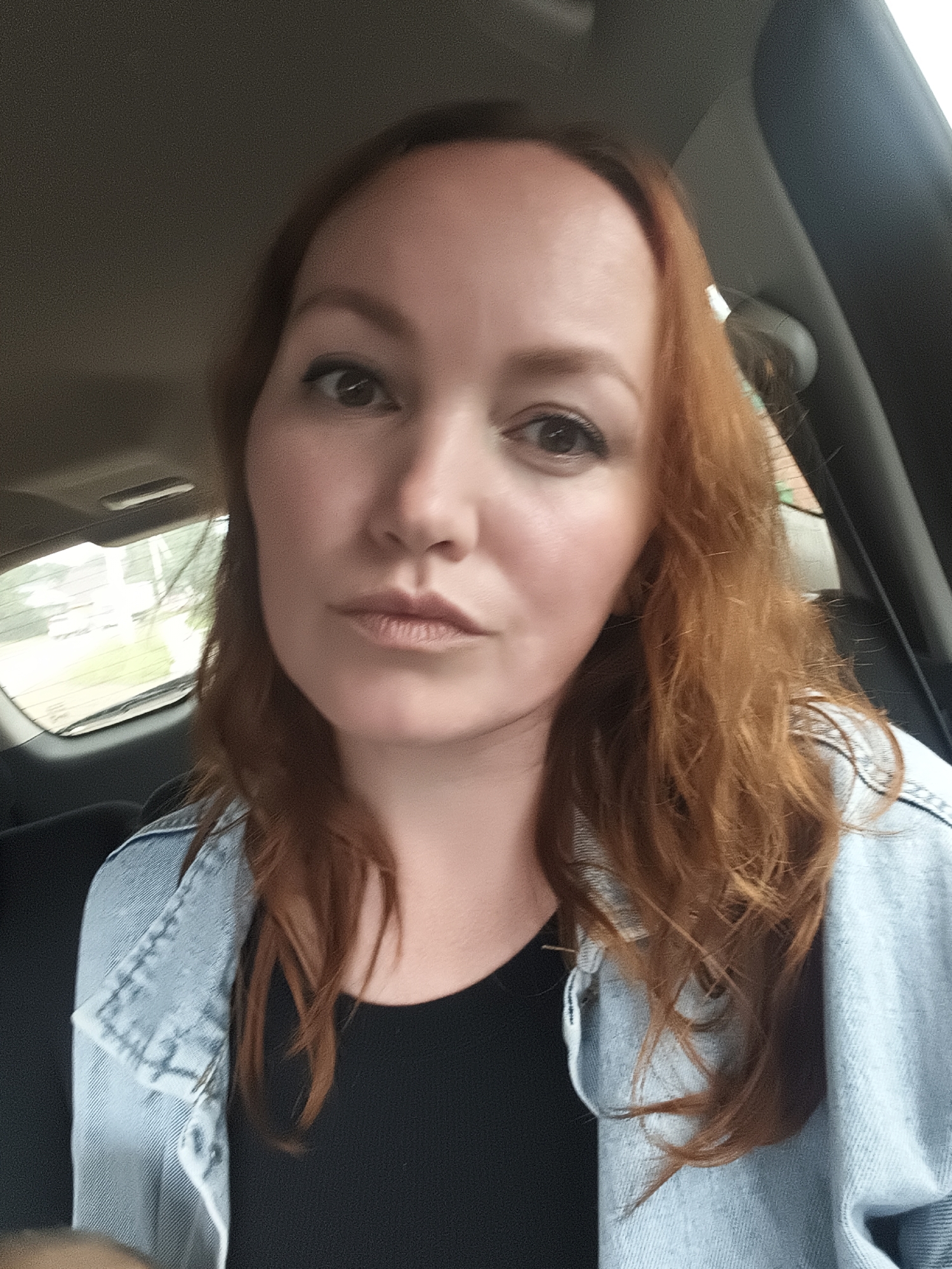 A selfie of a woman with auburn hair, wearing a light blue denim jacket over a black shirt, is taken inside a vehicle. The woman is looking directly at the camera with her mouth slightly open. The interior of the car is visible, with the headrest of the passenger seat behind her and a window showing a blurred outdoor scene of trees and buildings, consistent with a residential area in the city of Chaikovsky, Russia.