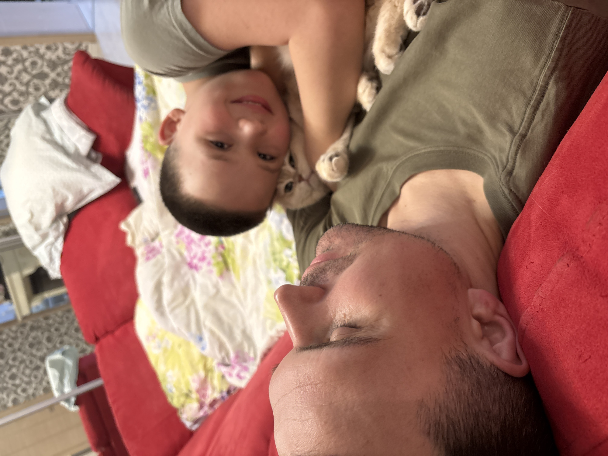 A selfie of a man and a boy in Chaikovsky, Russia. The man is lying on his side on a red couch and has close-cropped dark hair and stubble. His eyes are closed. A boy is lying on top of him with a cat nestled between them. The boy is wearing a green t-shirt and smiling. The cat is light-colored with blue eyes. A floral patterned pillow is nearby.