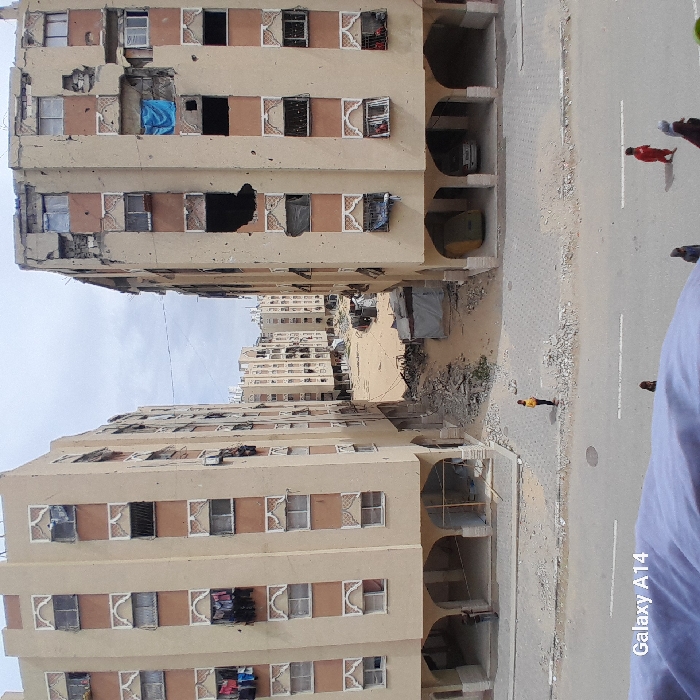 This image depicts a damaged urban streetscape in Khan Yunis, Palestinian Territory, under an overcast sky. Two multi-story residential buildings, constructed of light beige facades with reddish-brown decorative panels, frame a central, debris-strewn sandy clearing. The building on the left shows some wear and items like clothes hanging from windows, while the building on the right exhibits extensive structural damage, including numerous large holes in its facade, shattered windows, exposed interiors, and a partially collapsed roofline. A blue tarp covers a window opening on the damaged right building.
In the foreground, a paved street, partially covered in dirt and rubble, runs horizontally across the frame. Adjacent to the street, a damaged sidewalk area is visible, also littered with debris. One individual in a yellow top stands on this sidewalk, observing the scene. Several other individuals are walking on the paved street in the lower foreground, including one person wearing red. Under the arched ground-level openings of the right building, a yellow vehicle and a white car are parked. Beneath the left building's arches, two figures are partially visible. The central clearing between the buildings is filled with rubble, sand, and what appear to be makeshift shelters or debris piles, extending towards a further cluster of similarly styled, seemingly damaged buildings in the background.