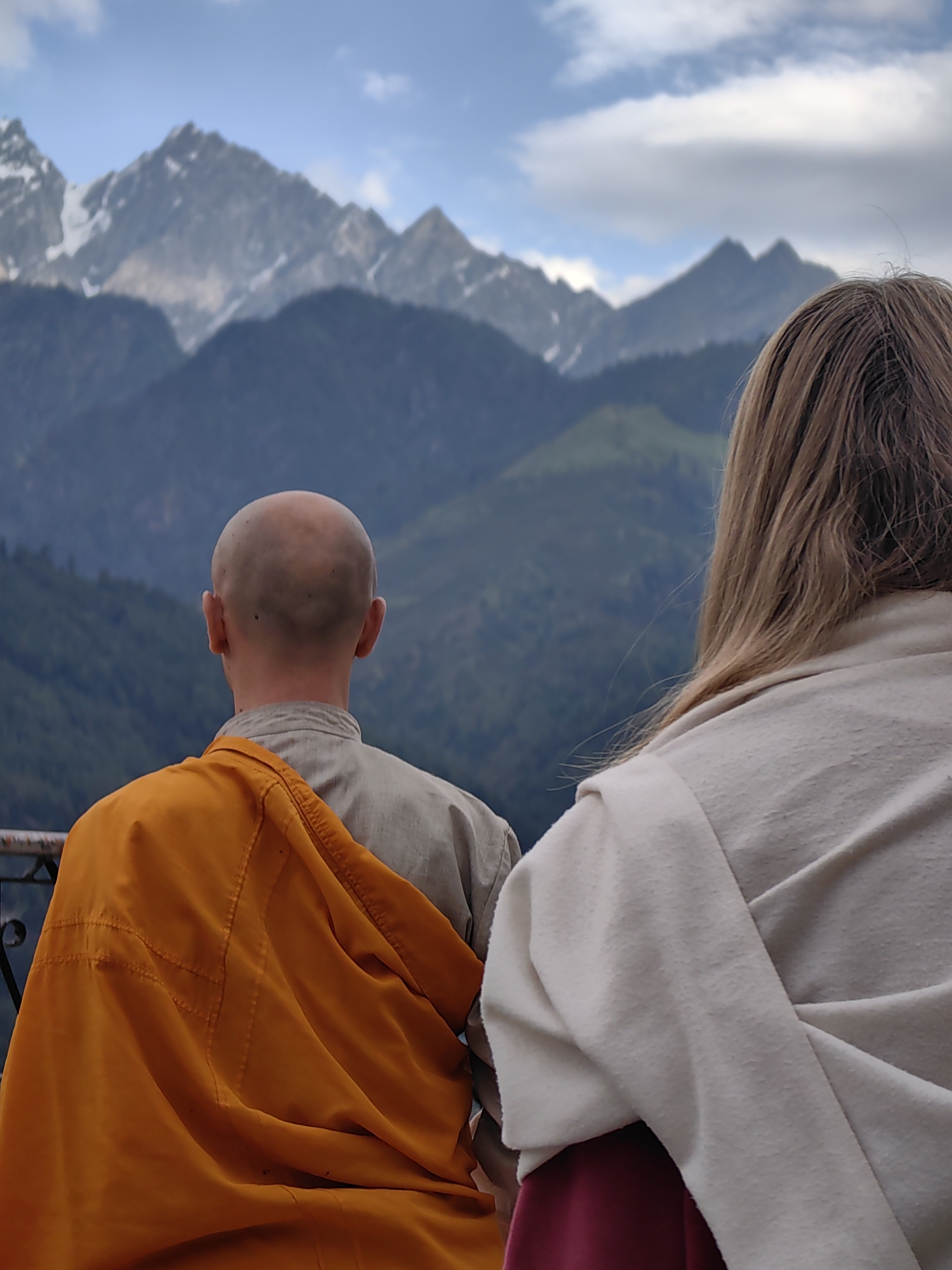 In Vashisht, India, two individuals are captured from behind, facing a majestic mountain range. The person on the left, appearing to be a monk, wears a beige robe with a vibrant orange sash draped over their shoulder. Their head is shaved. To their right, a person with long, blonde hair is seen wearing a light-colored wrap and a maroon garment underneath. The background features imposing mountains, some with snow-capped peaks, under a cloudy sky. The scene suggests a moment of contemplation or spiritual observation in a serene, natural setting.
