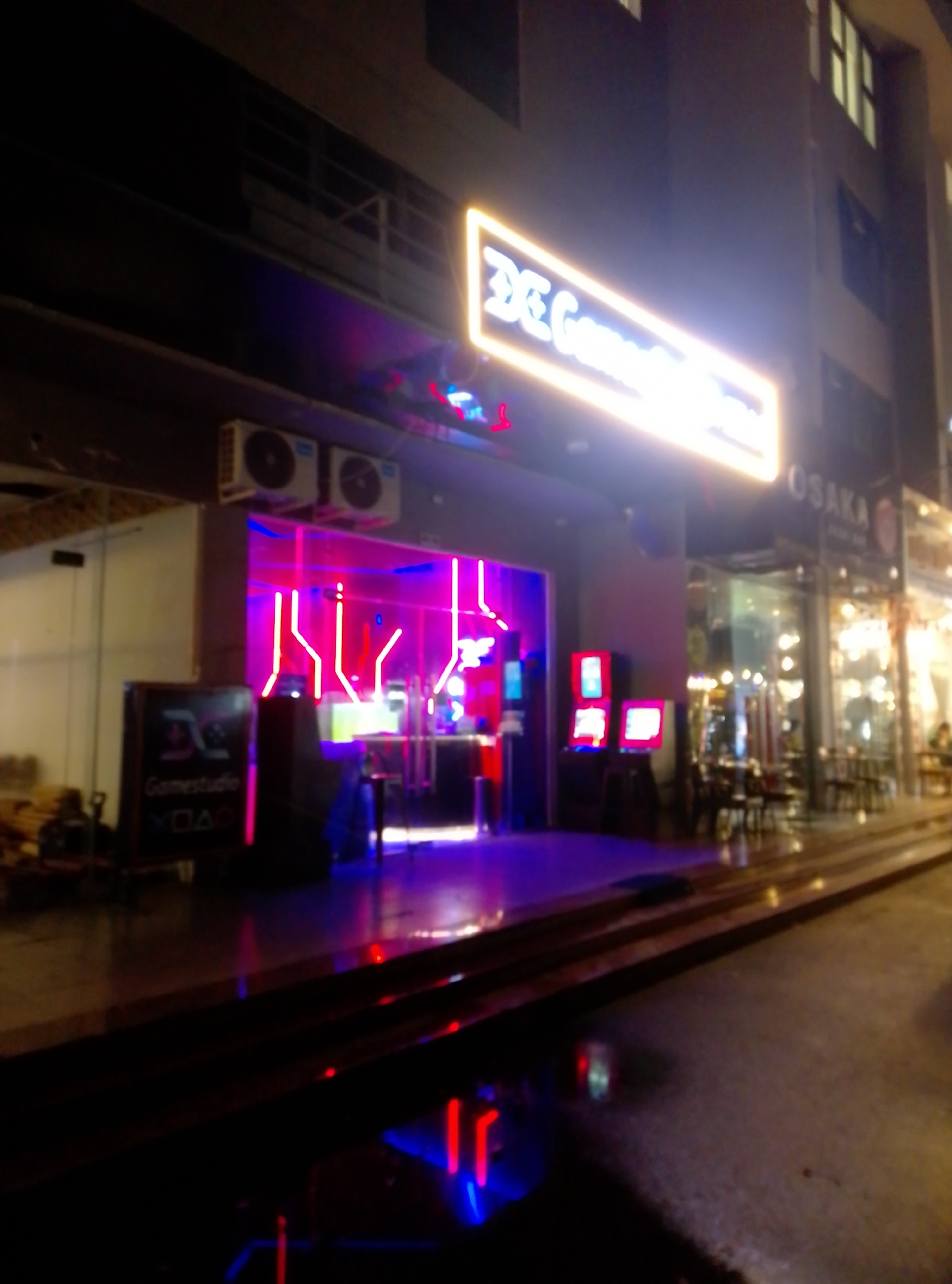 The image shows a nighttime view in Bac Nha Trang Ward, Vietnam. Visible is the exterior of what appears to be an entertainment venue, possibly a gaming center, indicated by the illuminated "DC Gamestudio" sign above the entrance. To the right of this venue is a restaurant or bar called "Osaka", also illuminated. The storefronts are adjacent to each other, with raised entrance platforms and steps leading up from the street. The street is dark and wet, reflecting the colored lights of the businesses. Several air conditioning units are installed above the "DC Gamestudio" entrance. Within the gaming studio, pink and blue neon lights are prominent. Several monitors are visible inside the venue. Outside of the venue, there is a gaming console with a screen in front of a sign. There are chairs and tables set up outside Osaka.
