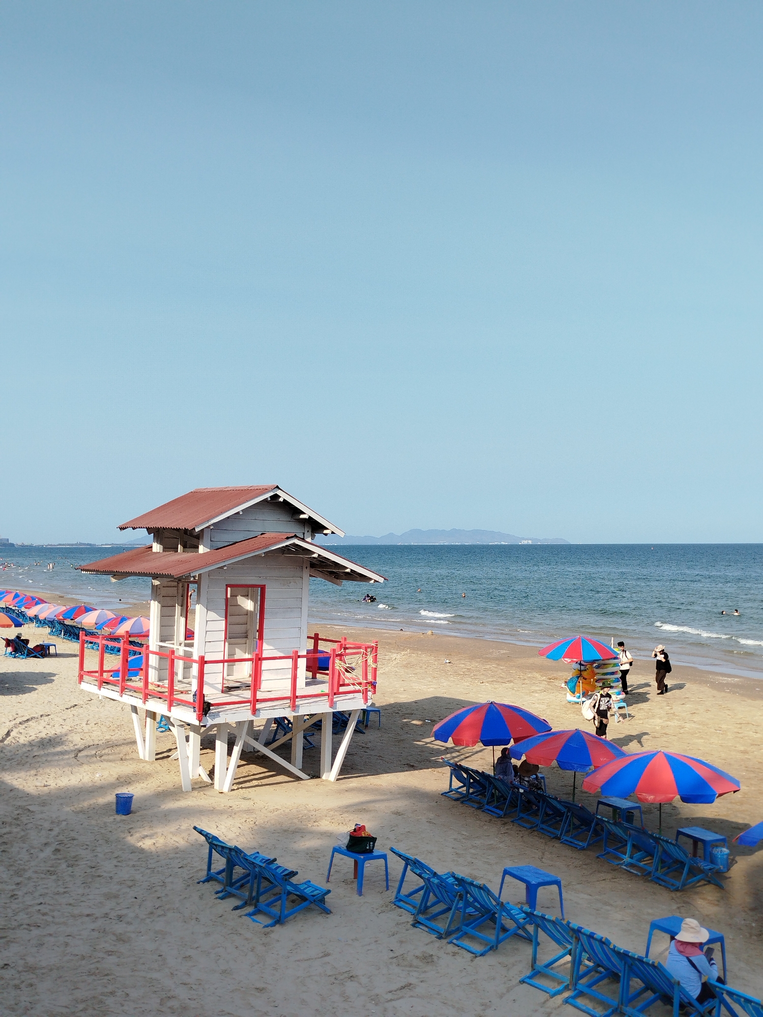 The image shows a beach scene with a lifeguard tower in the foreground. The tower is a two-story wooden structure painted white with a red roof and red railings. To the left of the tower, several rows of lounge chairs under colorful beach umbrellas stretch along the beach. In the water, several people are swimming. In the background, there is a coastline with mountains on the horizon. On the beach to the right of the tower, there are several beach umbrellas and people walking around. The sky is blue. While I cannot determine the precise location in Ho Chi Minh City based on the image alone, it is most likely a coastal area within the greater Ho Chi Minh City metropolitan area, or more generally, Vietnam.