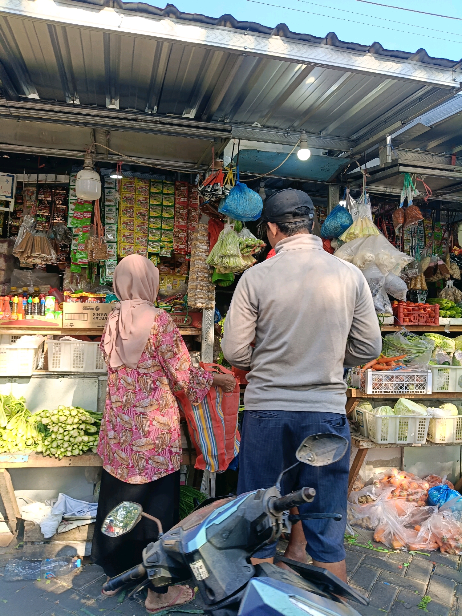 A street-side market stall in Surabaya, Indonesia, is depicted from behind. Two individuals, a man and a woman, are engaged in shopping. The woman, wearing a pink hijab and a patterned long-sleeved shirt over a black skirt, is holding a striped shopping bag. The man, wearing a grey long-sleeved shirt and dark blue shorts, stands beside her. The stall is laden with fresh produce, including leafy green vegetables in baskets and plastic bags, carrots, and what appear to be cabbages. Various packaged goods, such as instant noodles and spices, are displayed on shelves behind the vegetables. The stall is covered by a corrugated metal roof supported by a metal frame. A motorcycle is parked in the foreground, its side mirror reflecting the scene. The ground is paved with interlocking bricks and is littered with some debris. The lighting suggests it is daytime, with sunlight casting shadows on the ground.