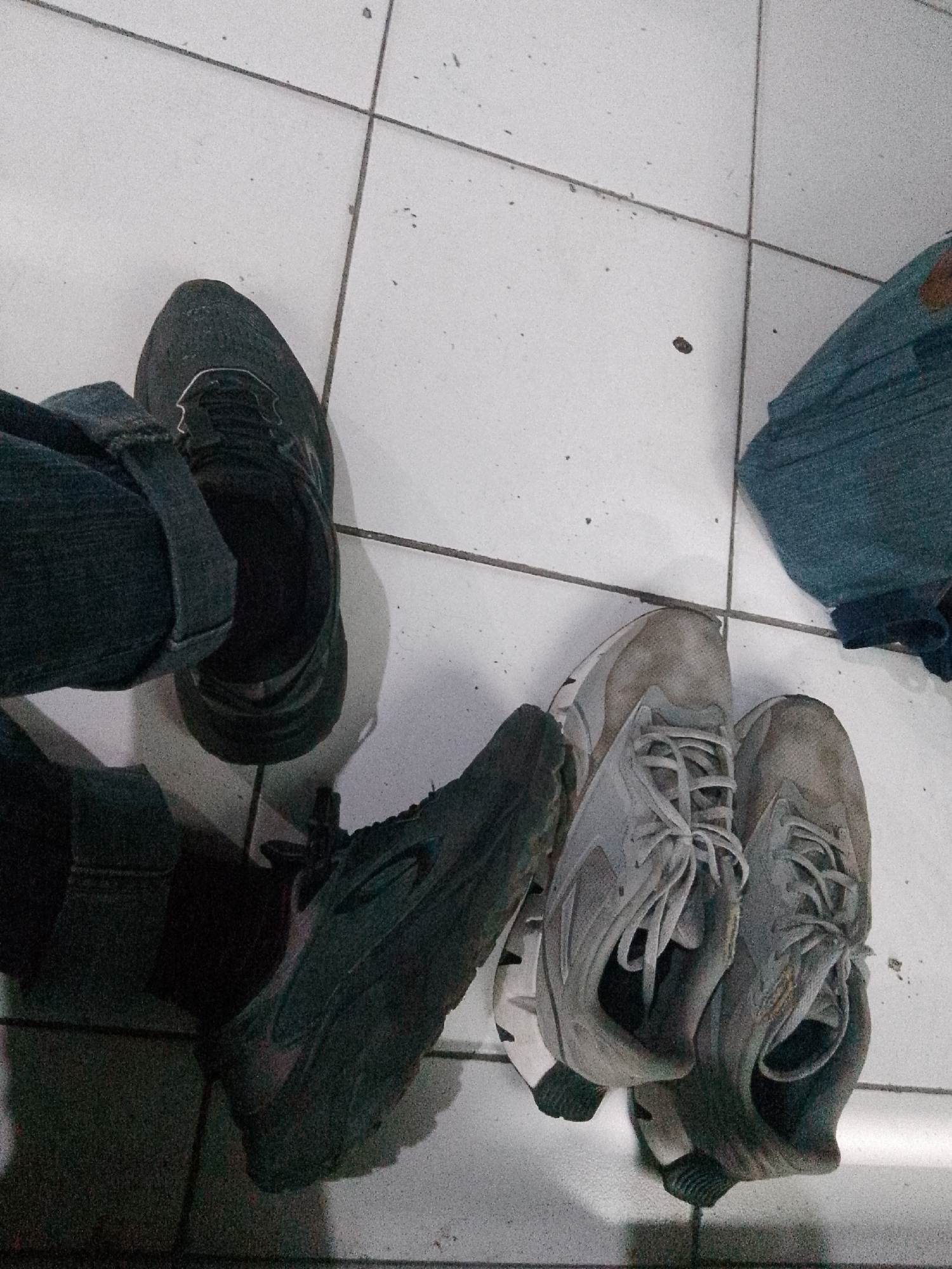 The image depicts a pair of black sneakers and a pair of light gray sneakers on a white tiled floor. A person's legs, clad in dark denim jeans, are visible on the left, wearing the black sneakers. To the right, the light gray sneakers are positioned next to a crumpled piece of blue fabric, possibly clothing. The lighting is dim, and the image appears to have been taken from a high angle. There is no discernible information in the image to indicate a specific location within the city of Trenggalek, Indonesia. The scene is static and shows only footwear and fabric.