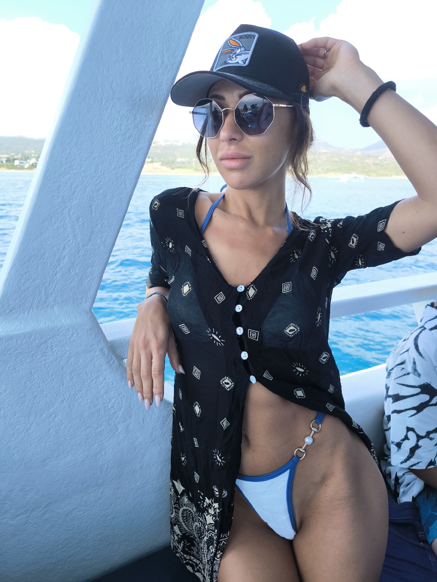 A woman with light brown hair, wearing a black patterned cover-up over a blue bikini top, a black baseball cap with a Bugs Bunny patch, and sunglasses, leans on a white railing on a boat. Her left hand rests on the railing while her right hand is raised to adjust her cap. The boat is on blue water, with a coastline and hills visible in the background, suggesting a location near Neo Chorio, Cyprus. The overall scene is bright and appears to be taken during the daytime, likely on a sunny day.