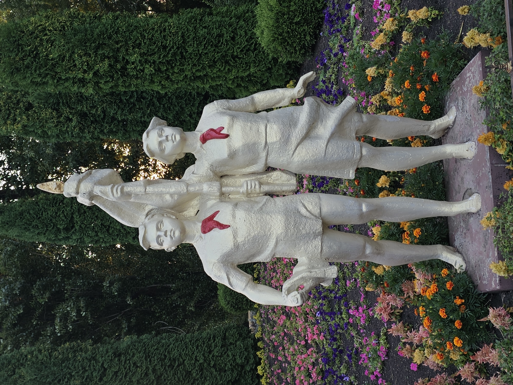 The image shows a statue of two young people, a boy and a girl, holding a flag pole. They are dressed in simple clothing and have red scarves around their necks. The statue is made of a light-colored material, possibly plaster or concrete, and is positioned in a garden with a variety of flowers in bloom. Behind the statue, there are tall green trees. The statue is located in Cherkessk, Russia, and is a monument dedicated to the Pioneers, a Soviet youth organization. The boy is on the left, holding a shoe in his left hand, while the girl is on the right. Both figures are looking forward with serious expressions. The flag pole is decorated with a golden tip. The flowers in the foreground are in shades of pink, purple, orange, and yellow, creating a colorful contrast with the statue and the greenery. The overall impression is one of youth, optimism, and dedication.