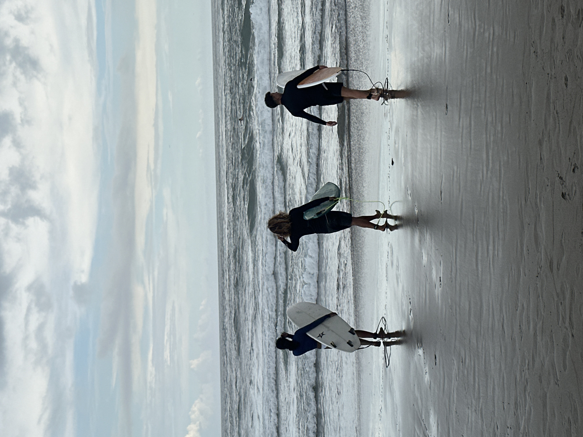 The image, oriented horizontally, depicts a serene coastal scene at a beach in North Kuta, Indonesia, under an overcast sky with diffused lighting. The wet sand in the foreground shows ripples and footprints, transitioning into the ocean which stretches across the midground and background with gentle, breaking waves.
Three individuals, presumably surfers, stand near the water's edge, each holding a surfboard and facing towards the ocean. They are all dressed in dark swimwear, likely rash guards or wetsuit tops and shorts.
On the left, one surfer with light, curly hair holds a light green surfboard, with a leash visible on their ankle. In the middle, another surfer holds a light-colored board with a darker bottom. On the right, the third surfer holds a white surfboard featuring a black star or cross design on its underside, with visible fins and a leash.
The individuals appear to be either preparing to enter the water for a surf session or observing the wave conditions. Their focused stances suggest anticipation or assessment of the ocean. The cloudy sky adds to the soft, even illumination of the scene. No discernible text is visible other than the design on one surfboard. The overall impression is one of calm readiness for a surfing activity.