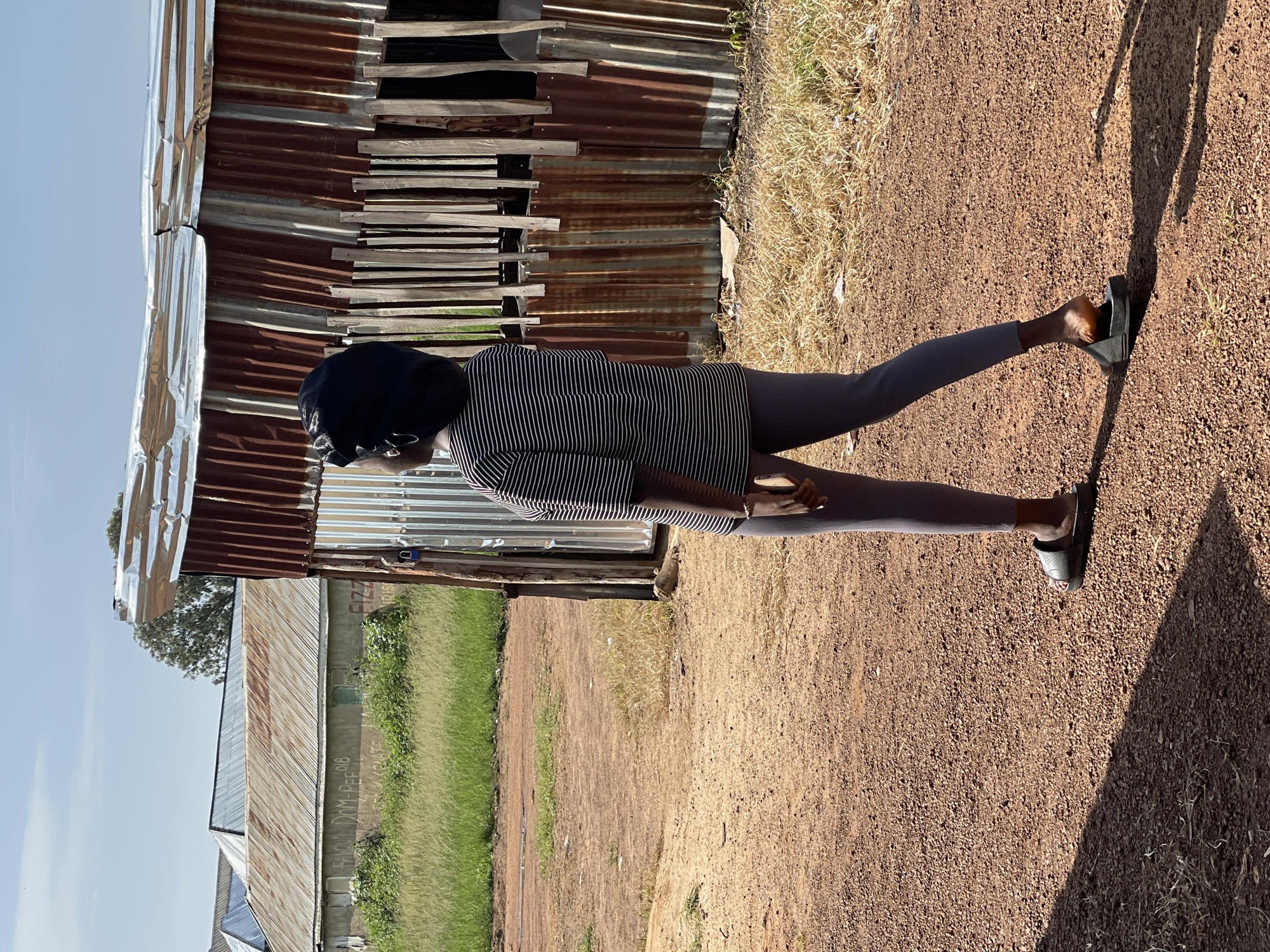 A young African female with dark skin, wearing a black headscarf, a black and white striped t-shirt, and gray leggings, walks on a dirt road in a sunny outdoor environment. She is facing away from the camera and looking to her right. Her right foot is slightly raised as she walks, and she is wearing black sandals. Behind her, to her right, is a dilapidated structure made of corrugated metal and wooden planks. To her left, in the background, is a grassy field and more buildings with corrugated metal roofs. The sky is clear and blue. The overall setting appears to be a rural or semi-urban area in Durumi, Nigeria.