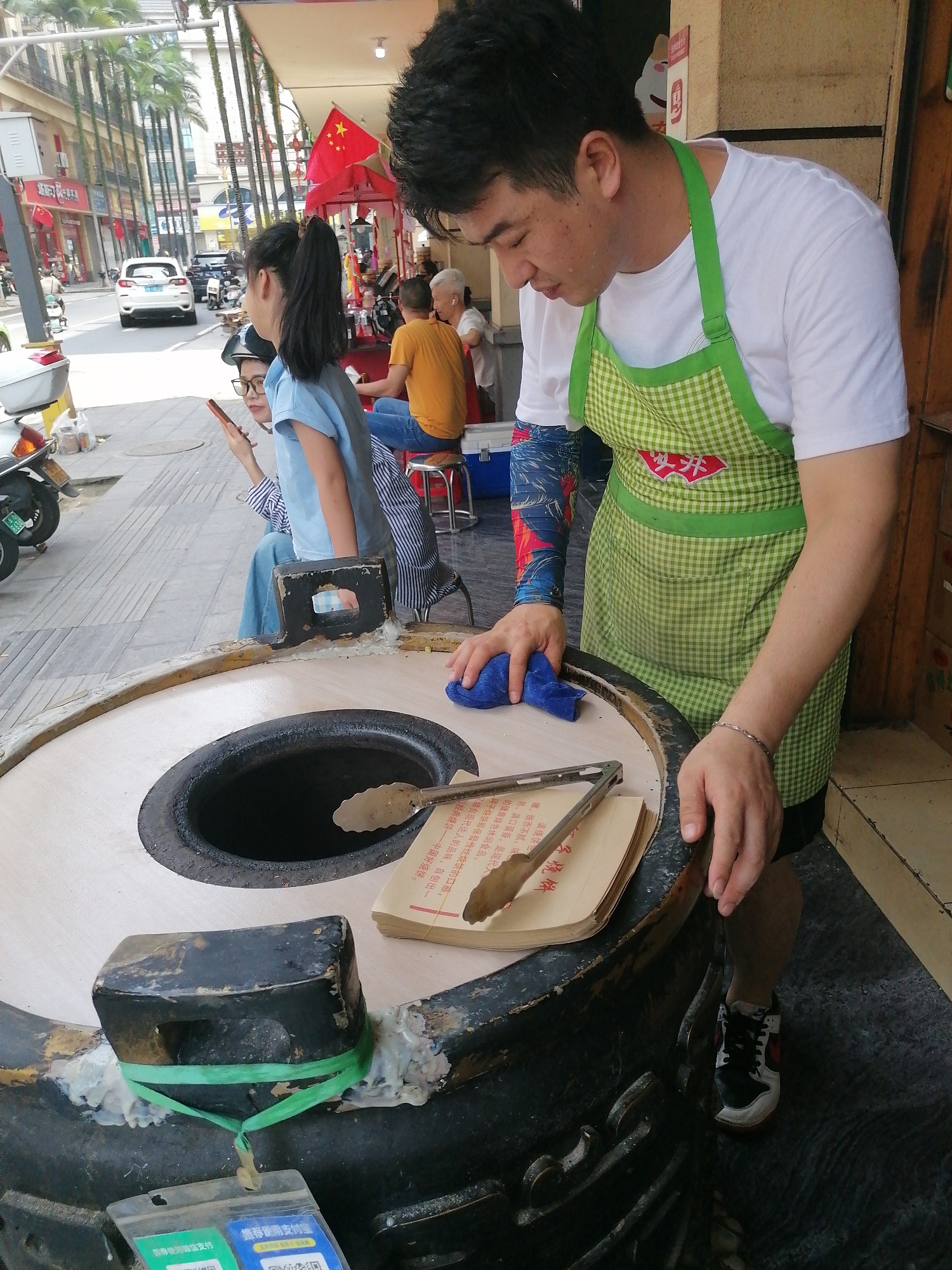 The scene captures a street food vendor in Hekou, China. A man wearing a white t-shirt and a green checkered apron is cleaning a large, circular oven with a blue cloth. The oven has a central hole, and beside it, there's a book with a pair of tongs resting on top. An object secured with a green band is on the oven’s edge, along with stickers showing QR codes for mobile payments. In the background, there are pedestrians seated at a roadside eatery, a Chinese flag, and a street with cars and palm trees lining the sides. A woman and a girl are seated on the sidewalk. The woman is holding a cellphone.