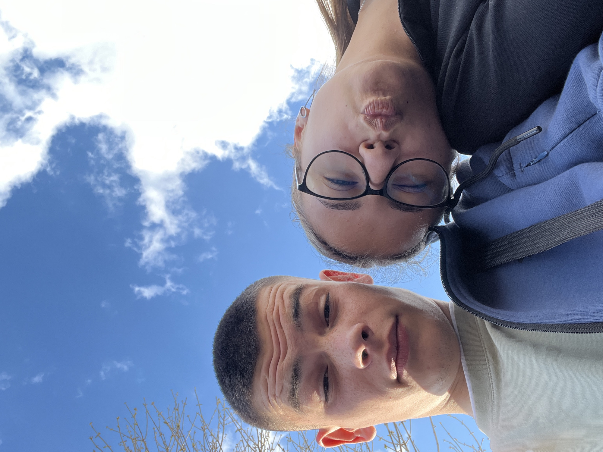 A low-angle selfie shows two individuals, a man and a woman, against a backdrop of a blue sky with scattered clouds. The woman, positioned at the top of the frame, wears glasses and has her eyes closed and is making a kissing face. She is wearing a black top and blue jacket. The man, positioned at the bottom of the frame, has short hair and is wearing a light-colored shirt. The bottom of the frame shows tree branches. The location of the image is unknown, but the prompt suggested Karavelovo, Bulgaria. There is no verifiable evidence of this.