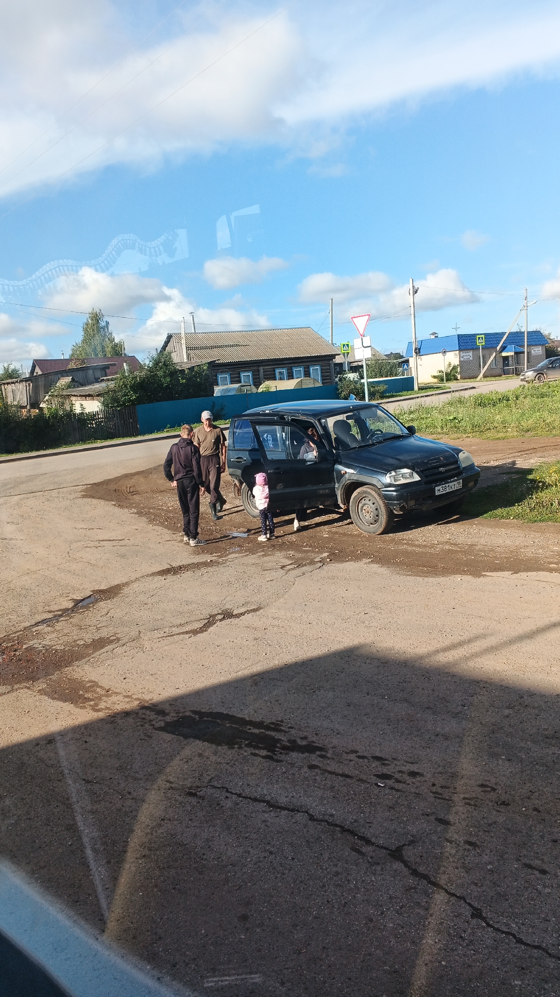 In the village of Kiyasovo, Russia, a dark-colored SUV is parked on a dirt road. The driver's side door is open, and two adults and a young child are standing near the vehicle. One adult, wearing a dark tracksuit, stands to the left of the SUV. Another adult, wearing a green t-shirt and a cap, stands to the right of the first adult. The child, wearing a pink hat and jacket, stands near the open SUV door. A building with a brown roof and wooden siding is visible in the background, along with other houses and trees. The sky is blue with scattered white clouds. The road is unpaved and appears to be wet in places. A road sign indicating a yield is visible in the distance. The scene suggests a pause in travel or a stop at a rural location.