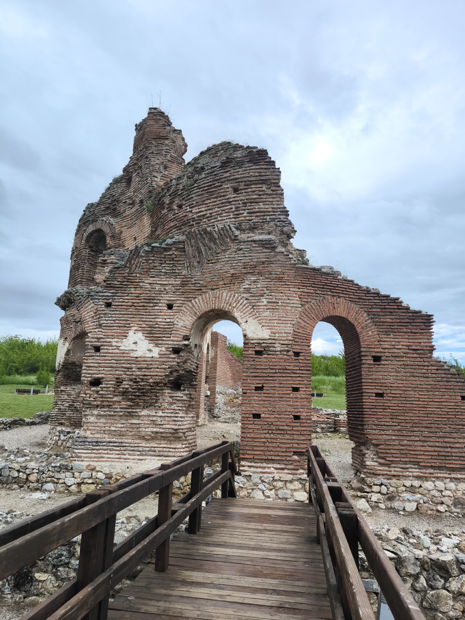 The image captures the remains of the Red Church, a partially ruined early Christian basilica located in Perushtitsa, Bulgaria. The primary subject is the dilapidated structure itself, constructed of red bricks and featuring arched openings. A wooden bridge spans a ground of rubble, providing a clear foreground element and leading the eye towards the ruins. The background features green vegetation, indicative of the natural surroundings. The sky is overcast, contributing to a somber and historical atmosphere. There are no visible people in the scene.