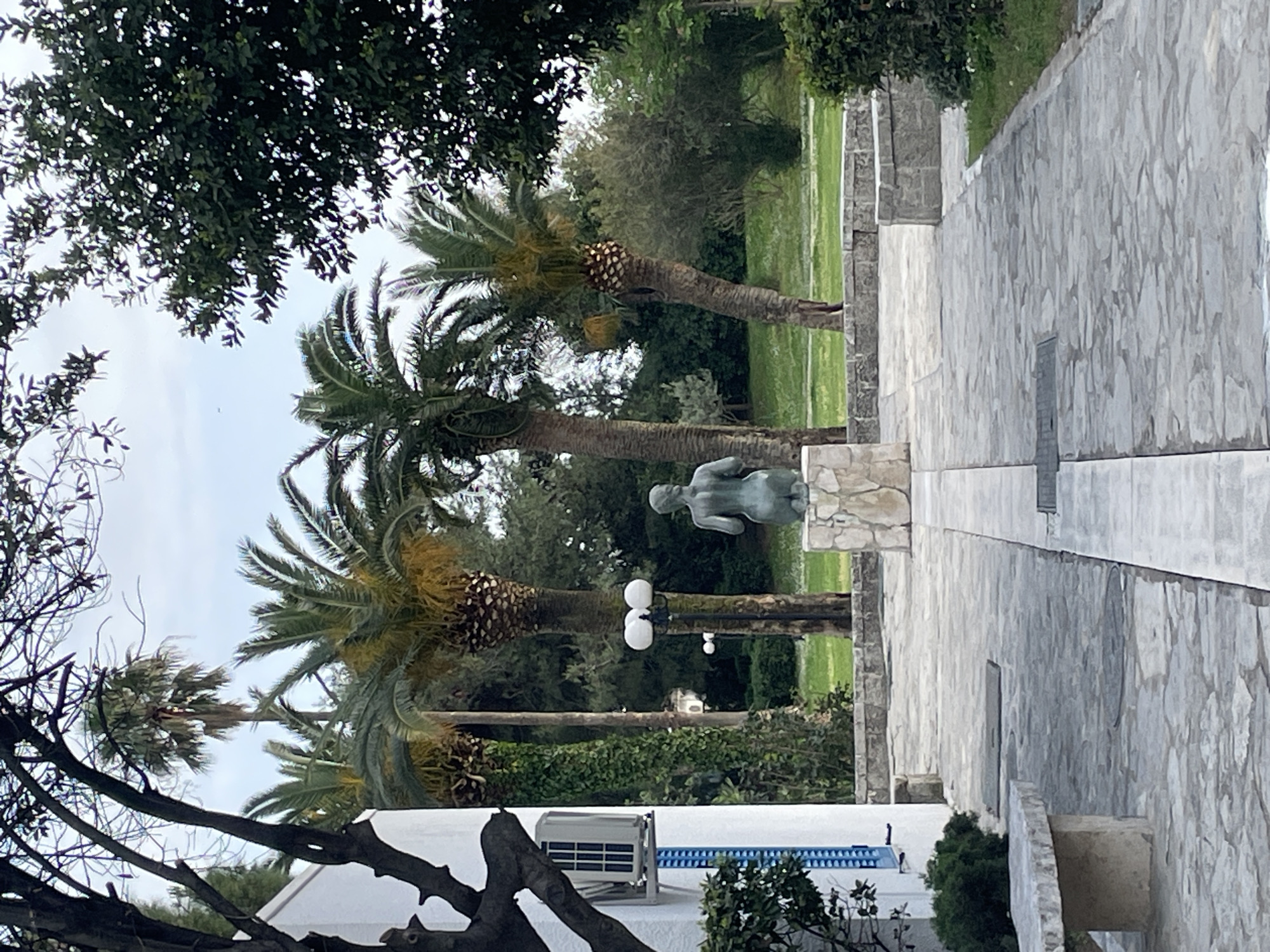 The image depicts a courtyard or park-like setting in Budva, Montenegro. Three palm trees are visible, aligned along a stone-paved walkway or terrace. A light green sculpture of a person is positioned near the center of the image. An air conditioning unit is visible on the side of a building below the sculpture. To the right is a light-grey stone wall and path. To the left is a partial view of building. The sky is overcast. The vegetation suggests a Mediterranean climate.