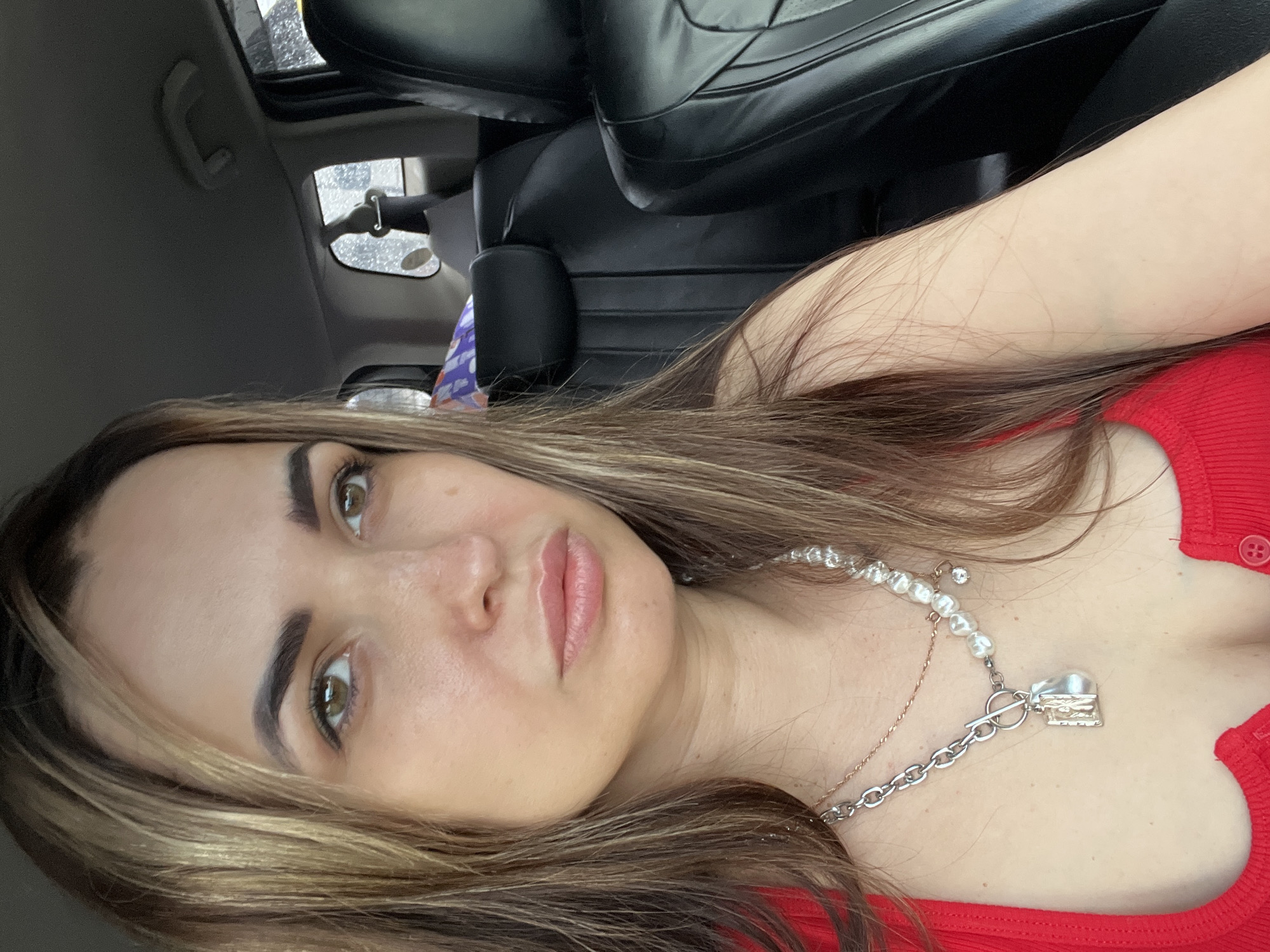 A close-up selfie shows a woman inside a car, captured from a slightly tilted, horizontal perspective. The woman has long, wavy light brown hair with subtle blonde highlights that frames her face and falls over her shoulders. She has distinct, dark eyebrows and light brown or hazel eyes that look directly at the camera. Her lips are a natural pink hue, appearing slightly parted. She is wearing a ribbed red top. Two layered necklaces adorn her neck: one is composed of white pearls with a small, clear charm, and the other is a silver chain with a toggle clasp and a small, detailed rectangular pendant that resembles a book or locket.
The background reveals the interior of a vehicle. Dark, possibly black, leather car seats are visible behind her. To the upper left, a car window shows visible raindrops, indicating rainy or wet weather. Portions of the car's interior ceiling and a rearview mirror can also be seen. The lighting suggests it is daytime, likely overcast due to the rain. The woman's expression is neutral and calm. There is no visible text in the image.
