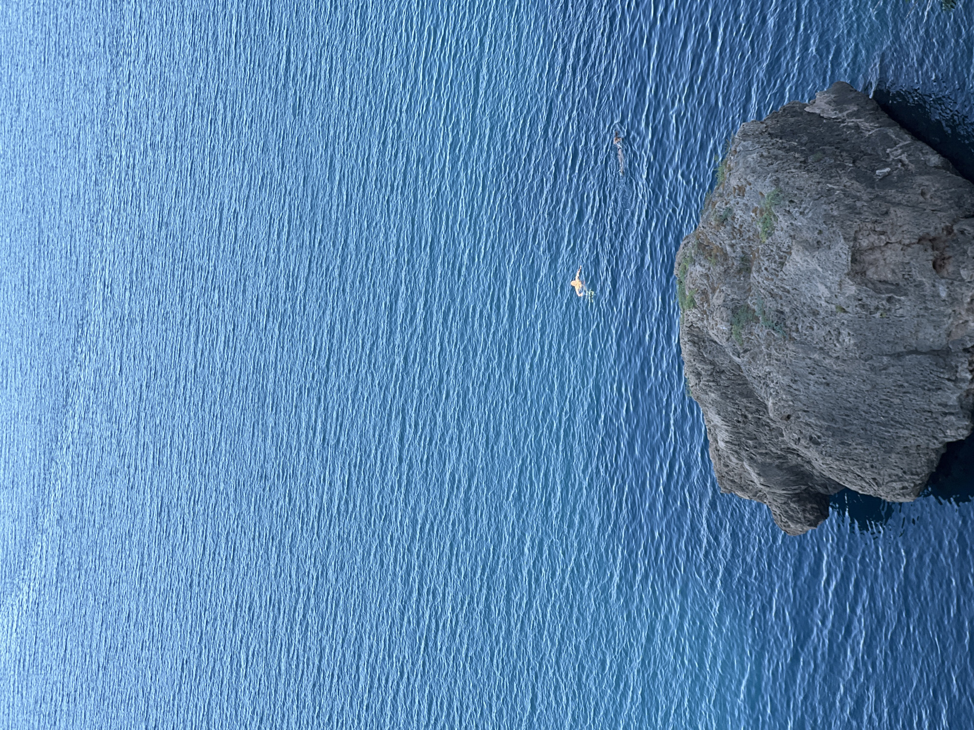 The image shows a rocky outcrop in the sea, with two swimmers in the water. The sea is a deep blue and appears calm with small ripples. The rock is large and grey, with some green vegetation growing on it. One swimmer is seen close to the rock, and another is further out in the water. The scene is likely a coastal area. This image could have been taken in Antalya, Turkey, given its geographical location known for its beautiful coastline and clear blue waters.