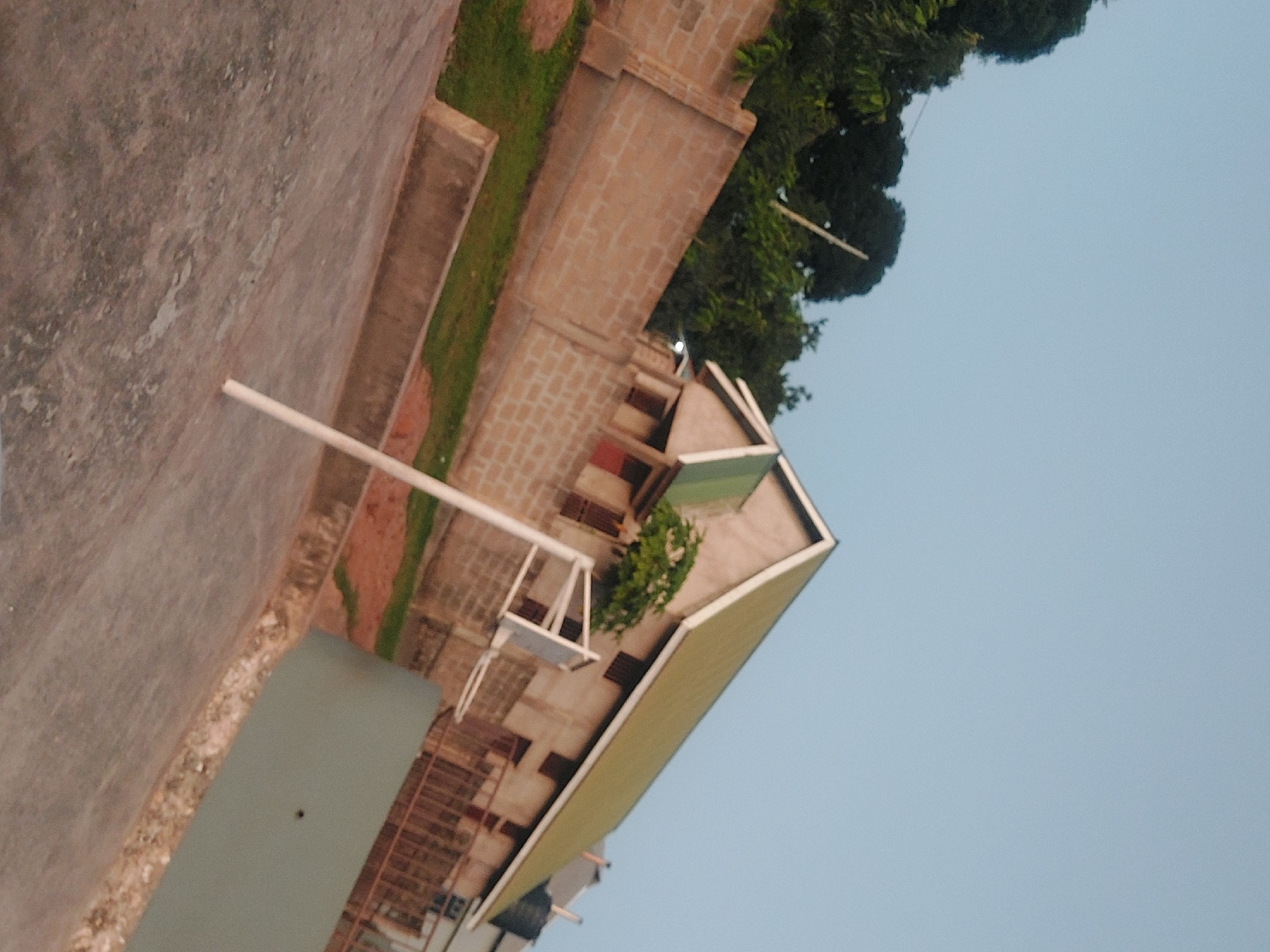 The image shows a building complex in Dar es Salaam, Tanzania. The primary structure is a multi-story brick building with a pale green roof. A swing set stands on a higher level of the complex. The sky is light blue. To the left is a concrete surface or walkway. Trees are located next to the building.