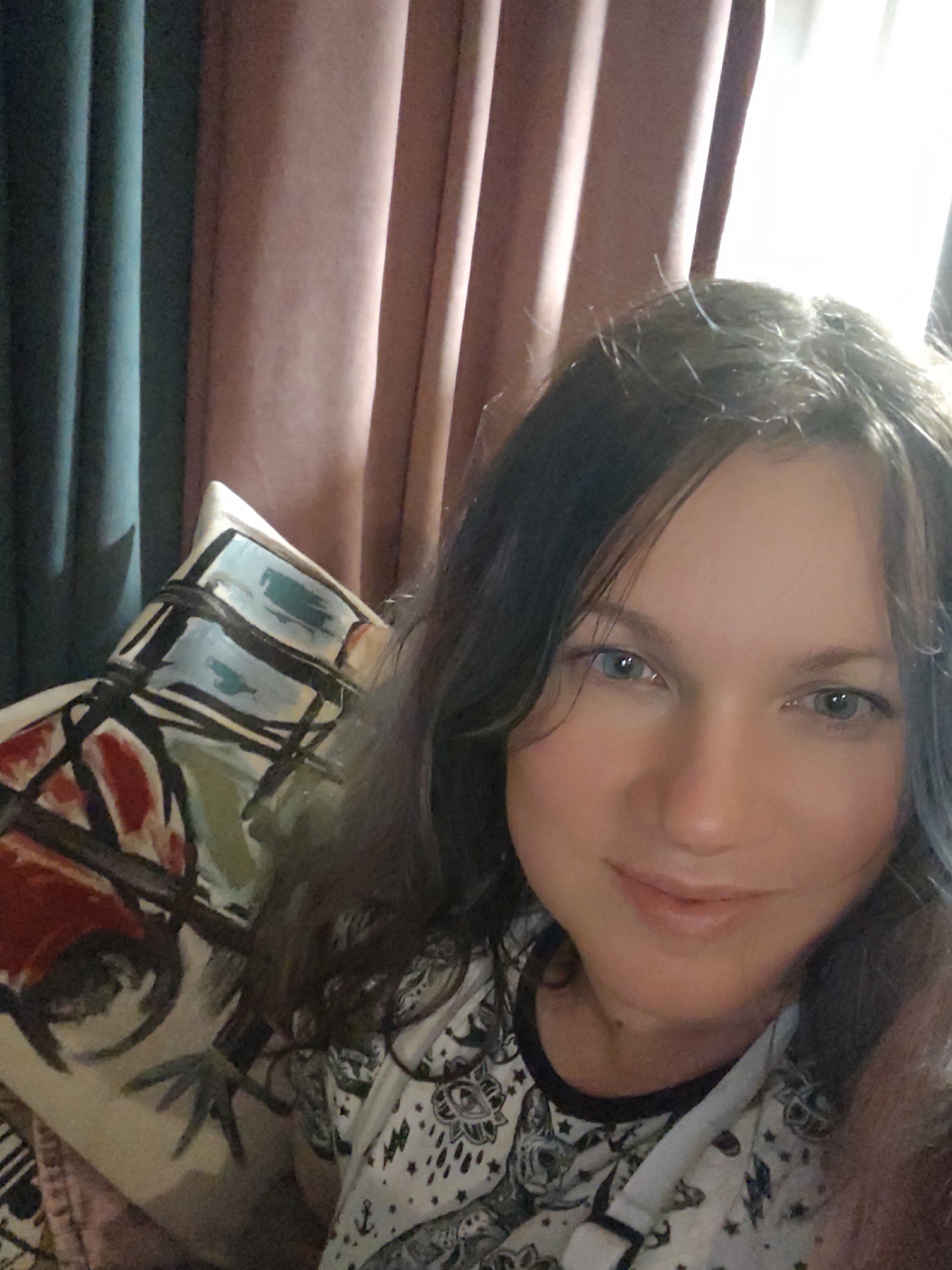 The image is a selfie taken by a woman with medium-length brown hair and blue eyes. She is smiling and looking at the camera. She is wearing a black and white t-shirt with a pattern of celestial bodies and animals. To her left, there is a pillow with an abstract design of red, blue, and black shapes. In the background, there are vertical curtains, one green and one pink, with light filtering through them from the right. The scene appears to be indoors, likely in a home. The location is identified as Свистуха (кузяевский С/о), Russia, which is a rural settlement.