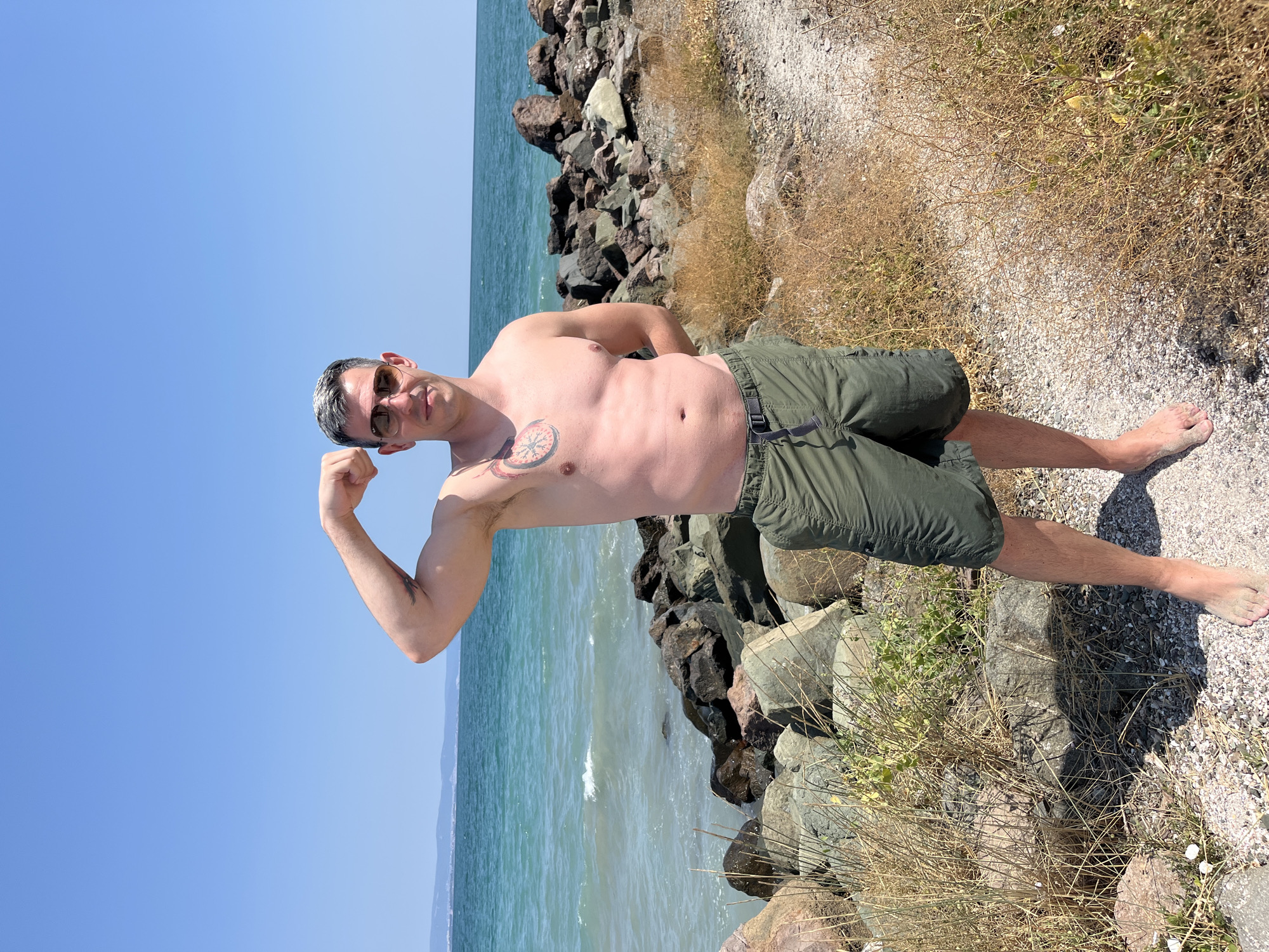 A man with grey hair and sunglasses stands on a rocky beach near the sea, flexing his bicep. He is wearing green cargo shorts and has a tattoo on his chest. The water is a vibrant turquoise, and the sky is clear blue. In the background, a faint coastline is visible. The foreground features dry grass and large rocks. The location is identified as Pomorie, Bulgaria.