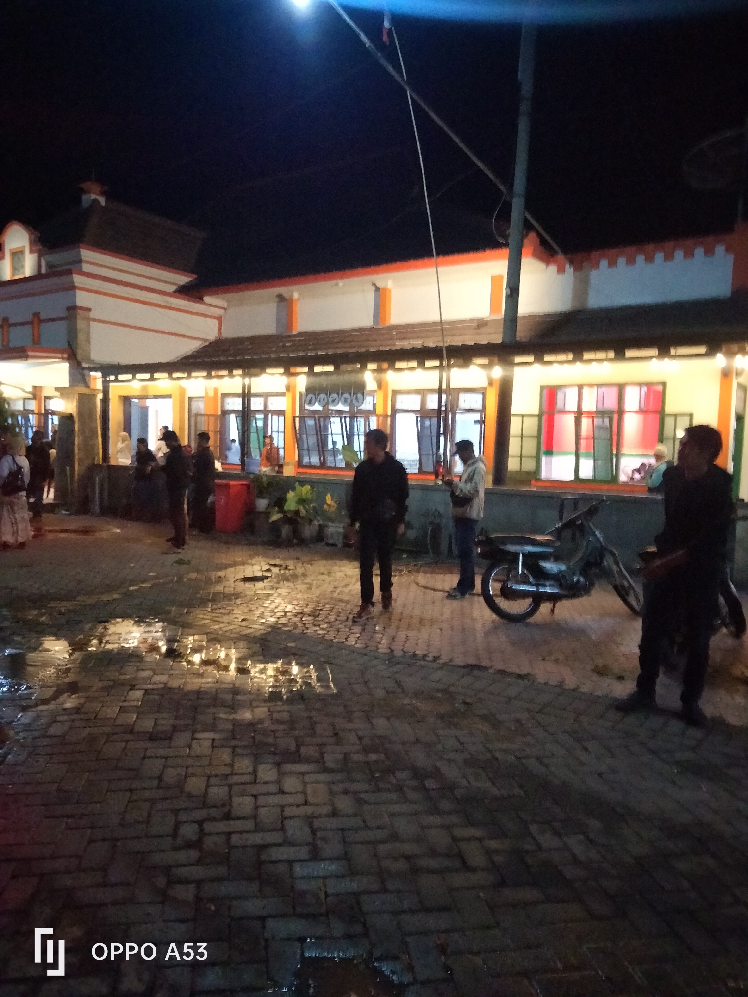 The image shows a group of people standing outside a building at night. The building has an orange and white facade with numerous windows. Some of the windows have colorful panes. The ground is paved with interlocking bricks and appears wet, reflecting the lights from the building. A motorcycle is parked to the right of the frame. A street lamp is visible in the upper portion of the image. The scene is consistent with an outdoor area in Buduan, Indonesia.