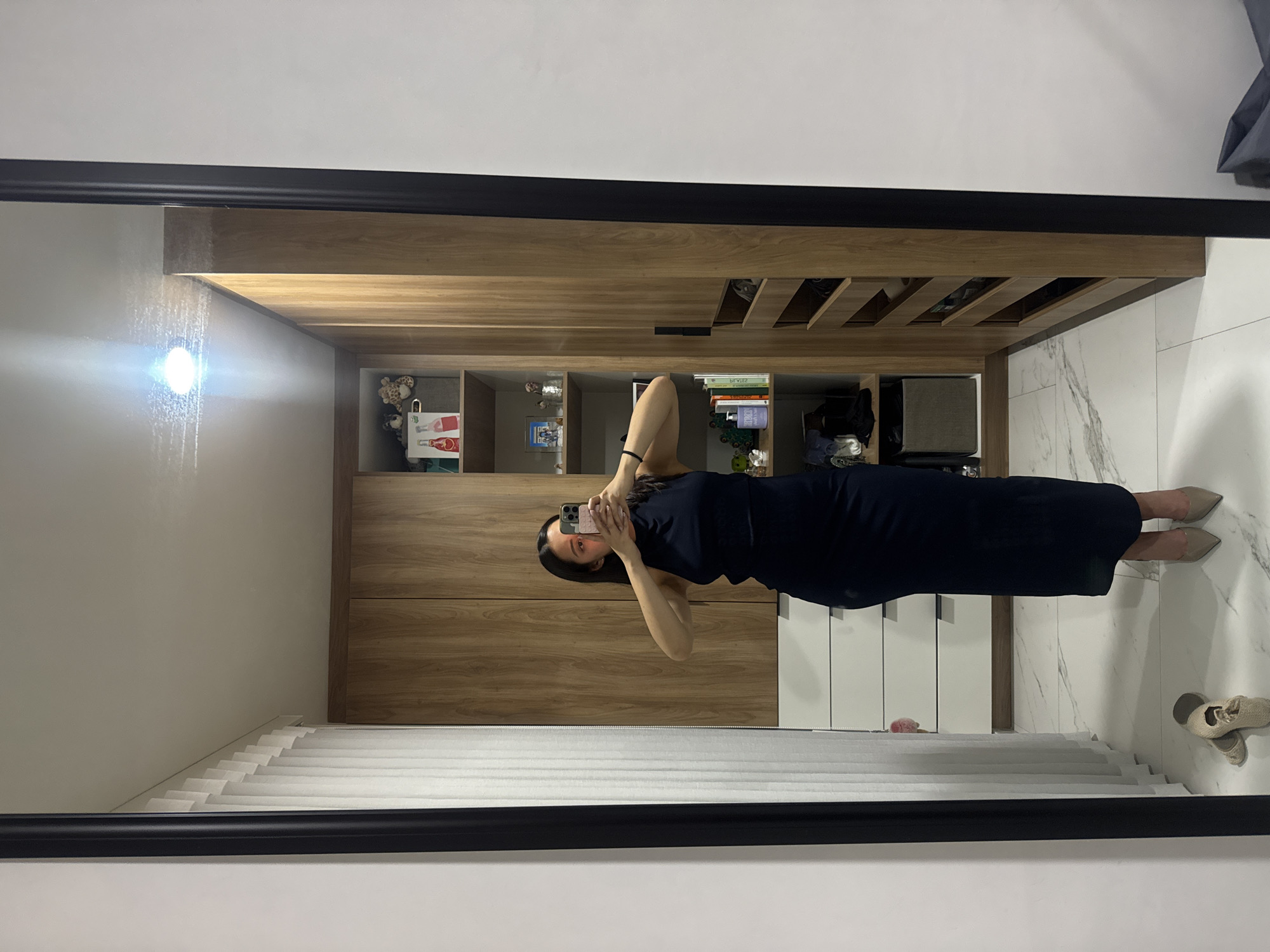 A person is standing in front of a mirror and taking a photo of themselves with their phone. The person is wearing a dark blue dress and beige shoes. The background includes a wall with a built-in wooden shelving unit and white marble flooring. The room also contains white blinds. No information indicates the location of the city of Puebla, Mexico.
