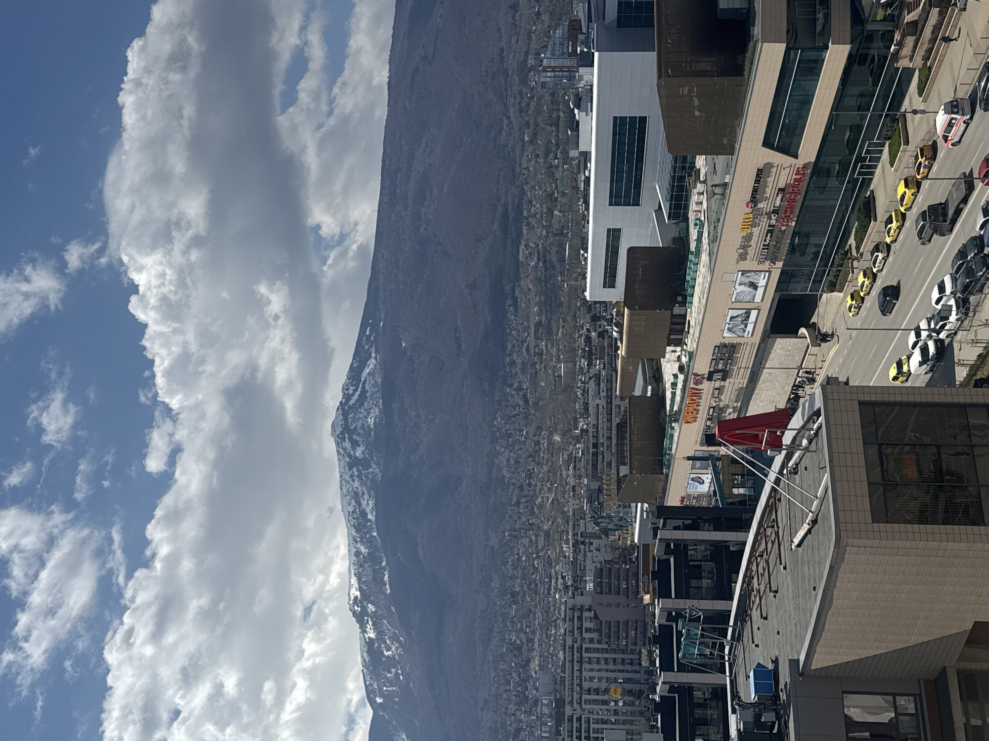 A daytime aerial shot of Sofia, Bulgaria, shows a mountain partially obscured by clouds on the left. Adjacent to the mountain, urban structures and buildings fill the scene, including a large shopping center visible on the right. A street runs alongside the building with parked and moving cars. The foreground features rooftops of various buildings.