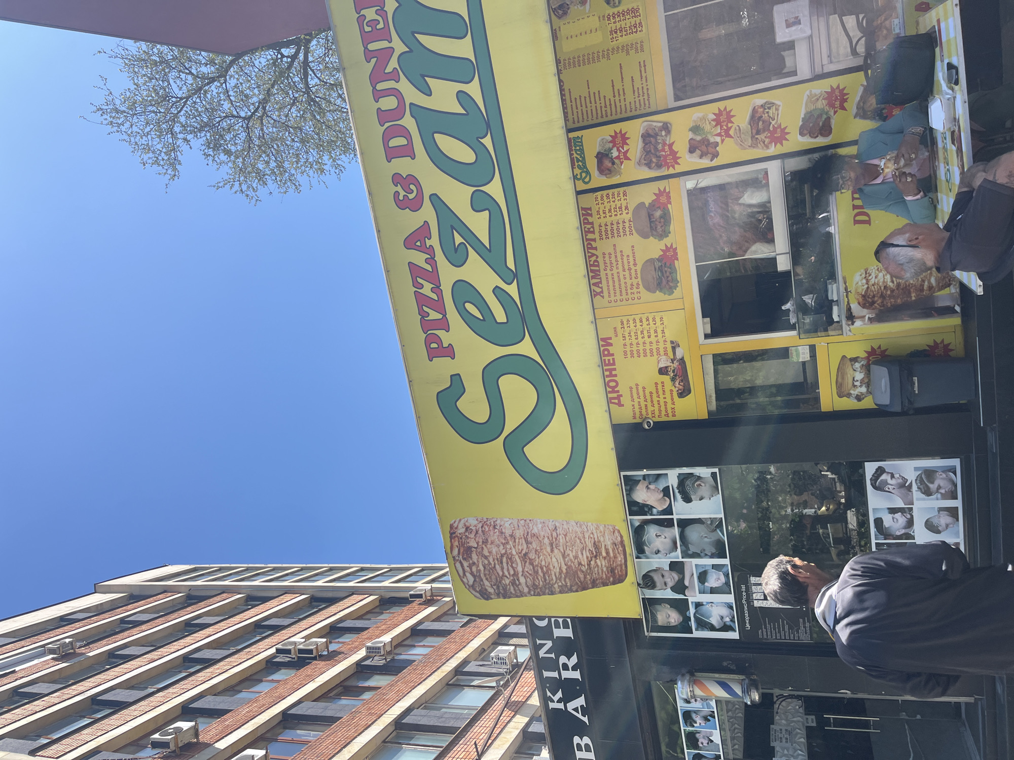 A photograph captures a street scene in Karnobat, Bulgaria, with a clear, blue sky overhead. A multi-story building with horizontally patterned windows occupies the left side of the frame. Dominating the mid-ground is a yellow-and-green sign for "Sezam Pizza & Duner," advertising food items in Bulgarian, with prices listed. Next to the food stand is King Barbers, with photographs of hairstyles adorning its facade. Two people are present near the food stand; one is dressed in a gray jacket.