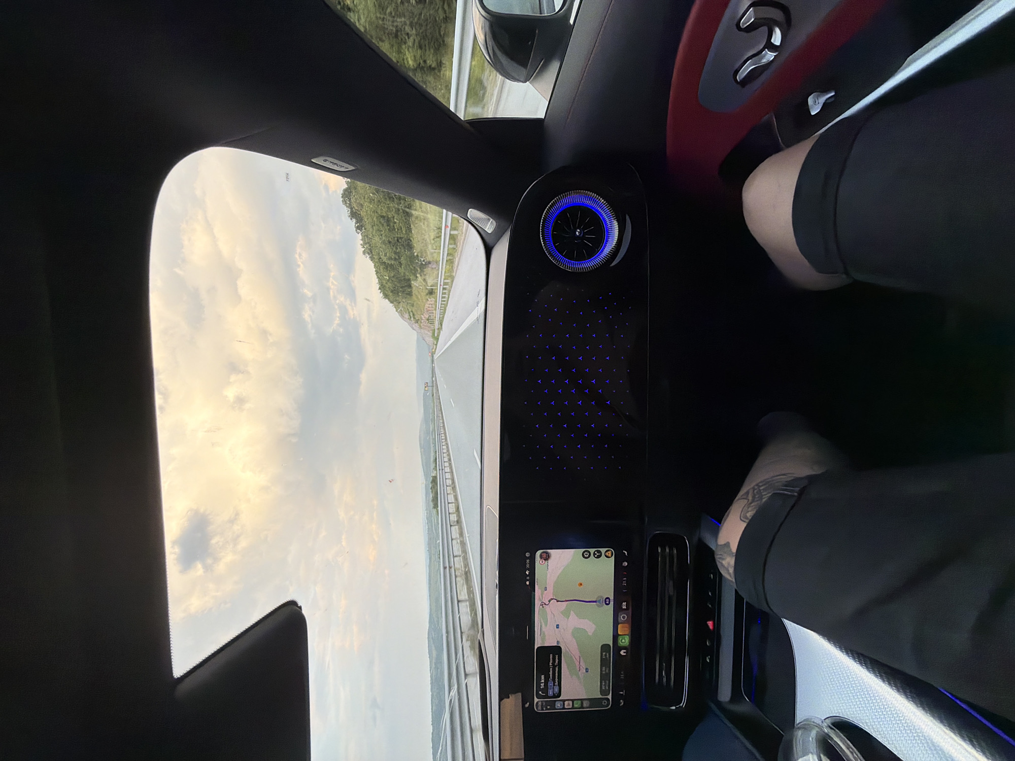 The image presents a detailed interior view from the front passenger seat of a modern vehicle, specifically a Mercedes-Benz, traveling on a multi-lane highway near Osikovitsa, Bulgaria. The perspective looks out through the windshield towards the road and the surrounding landscape.
In the foreground, two legs belonging to a person wearing black shorts are visible; the left leg exhibits a tattoo on the thigh. The car's dashboard prominently features two large digital displays. The left screen displays a navigation map, indicating a remaining distance of "14 km" and a speed limit of "140", with some text in Bulgarian. The right screen shows a decorative ambient lighting pattern consisting of multiple blue Mercedes-Benz star logos. To the far right of the dashboard, a circular air vent is illuminated with a bright blue ring. A section of the car's interior door panel, featuring red trim and silver seat adjustment controls, is visible on the right side. Overhead, a sun visor and the A-pillar with an "AIRBAG" label are partially visible.
Outside the vehicle, the highway stretches straight into the distance, flanked by metal guardrails on both sides. Rolling hills densely covered with green trees and foliage are visible on either side of the road, particularly prominent on the right. The sky is overcast with patches of brighter, warmer light, suggesting late afternoon or early evening. Small specks, likely dust or debris, are visible on the windshield. The vehicle is in motion, actively navigating a route.