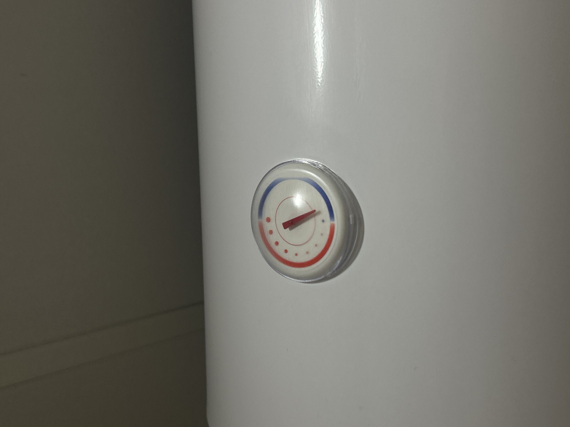 The scene, an interior view from Zlatograd, Bulgaria, displays a circular analog gauge embedded in a horizontal, white, smooth surface, likely part of a domestic appliance. The gauge, covered by a clear, slightly convex lens, features a white dial with a red dotted scale and crescent arc on the right, indicating warmer settings, and a blue dotted scale and crescent arc on the left, indicating cooler settings. A red indicator needle is positioned within the blue segment, pointing approximately to the 8 o'clock position. The upper portion of the image shows a uniformly dark, featureless background, possibly a wall or ceiling, under dim lighting conditions. No individuals or discernible actions are present.