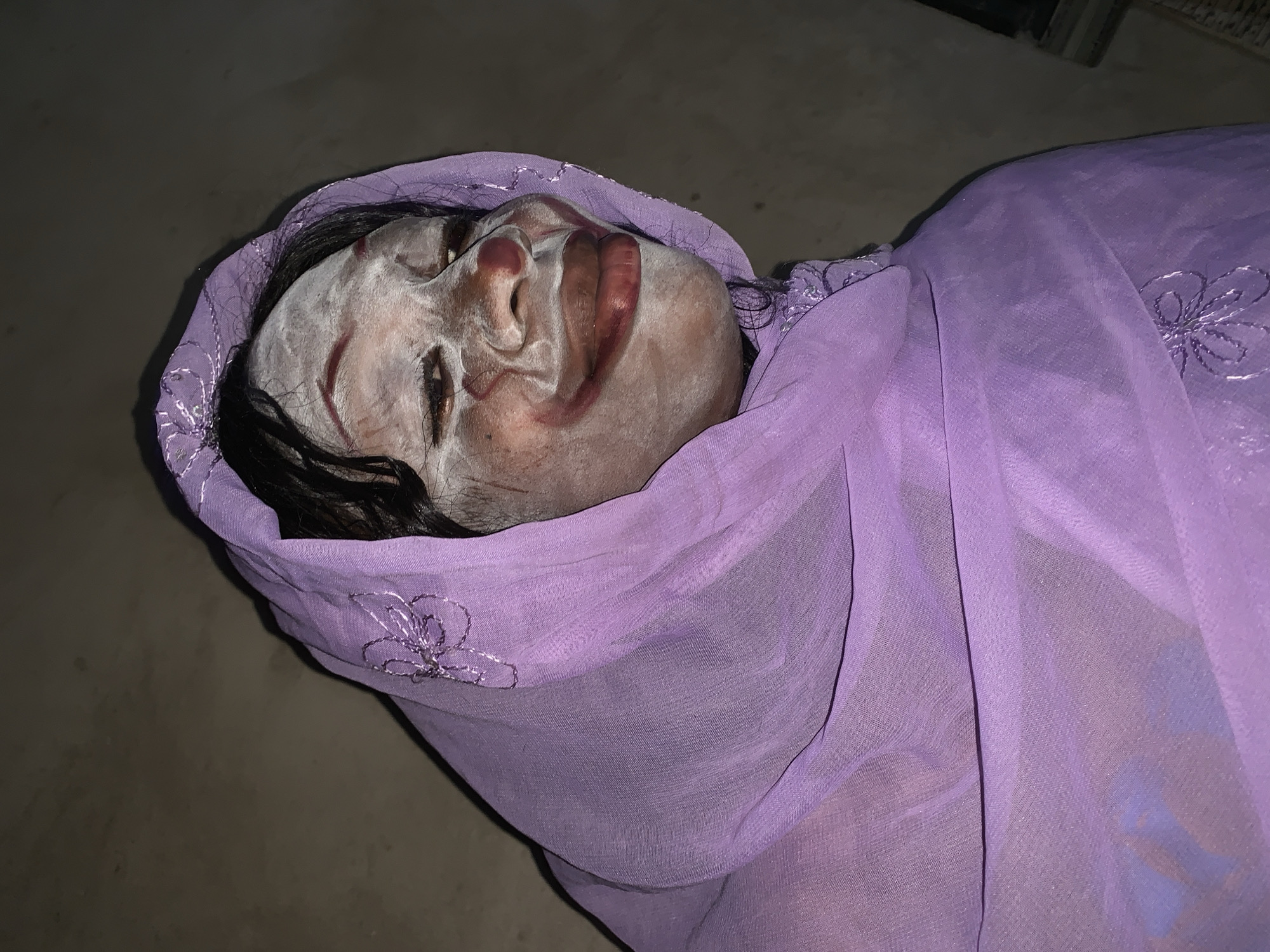 A deceased individual, possibly female, lies on a gray surface. The face is covered in a white substance. The person is draped in a sheer, light purple cloth embroidered with silver floral designs. The location is unknown but is stated to be Agban, Nigeria. The scene appears to be indoors, with limited ambient lighting. No further details about the immediate environment are discernible from the image.