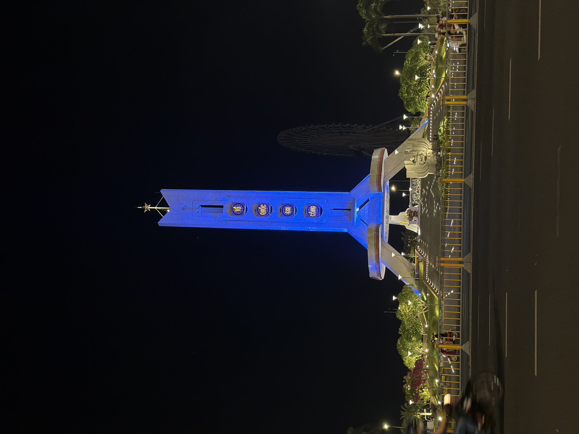 The image depicts a nighttime scene in Đà Nẵng, Vietnam, featuring a prominent illuminated monument. The monument is a tall, rectangular structure with a star-like finial at its apex, bathed in a vibrant blue light. Four circular emblems, each containing a symbol and text, are visible on the front of the monument. The text on these emblems appears to be in Vietnamese.
The monument is situated in a landscaped area with trees and flowering plants, illuminated by numerous streetlights. A yellow barrier separates the monument area from an adjacent road. The road has visible lane markings, suggesting it is a main thoroughfare. In the background, a large Ferris wheel is partially visible against the dark night sky, indicating a recreational or entertainment area nearby.
Several people are present in the scene, with some appearing to be walking near the monument and others further back near the trees and road. A few motorbikes are also parked or moving on the road. The overall atmosphere is one of a lively urban night scene. The image is taken from a low angle, emphasizing the scale of the monument. The dark sky indicates that it is nighttime, and there are no visible weather conditions to note.