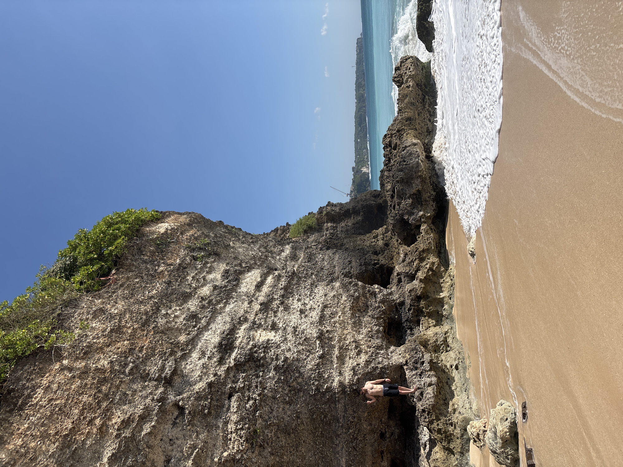 This image depicts a sunny beach scene in Kuta Selatan, Indonesia, featuring a large rock cliff on the left, a sandy shore, and the ocean.
On the left, a massive, textured rock cliff, primarily grey and light brown, rises prominently, with green foliage growing densely along its upper edge and sparsely in crevices. At the base of the cliff, several dark, irregularly shaped rock formations are visible, some forming shallow overhangs or small cave-like features.
Standing on these rocks at the base of the cliff, towards the lower-left corner, is a shirtless male individual. He wears dark shorts and a necklace. His left hand is raised near his face, possibly holding an object or adjusting something. He faces slightly right.
The foreground and mid-ground consist of a wide expanse of golden-brown sand, with clear patterns left by receding waves. Scattered dark rocks are visible on the sand and at the water's edge.
To the right, the ocean displays a gradient of blue, transitioning from a light turquoise near the shore to a darker blue further out. White foamy waves are actively breaking and receding onto the sandy beach. In the distance, a green, vegetated coastline is visible across the water, featuring several discernible structures, including what appear to be construction cranes and buildings.
The sky is a clear, bright blue, occupying the upper right portion of the image, with no visible clouds. The scene is well-lit, indicating bright daylight.