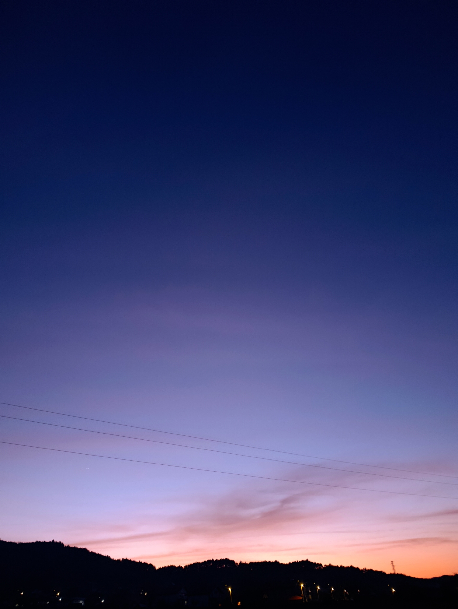 The image shows a dusk or dawn scene in Shiroko pole, Bulgaria. The sky transitions from a deep blue at the top to a gradient of purple, pink, and orange near the horizon. A few thin, horizontal wires stretch across the lower portion of the sky. Silhouetted against the horizon is a line of trees or hills, with scattered lights suggesting a town or village. A pole is visible on the horizon in the right side of the image.