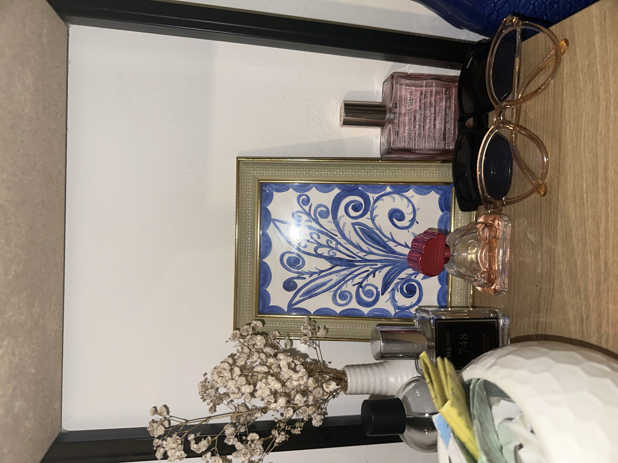 The image, oriented vertically, displays an indoor scene in Davao City, Philippines, featuring an arrangement of personal care items and decorative objects on a light wooden surface against a white wall. No people or specific activities are present.
At the center is a framed picture with a gold-colored frame, containing a vibrant blue and white pattern reminiscent of traditional ceramic or tile designs. To the right of the frame, near the top, rests a clear perfume bottle with a pink liquid and a silver sprayer. Visible text on this bottle includes "MULTIFUNCIONES NOSTRO CUERP26," "HUILE SECCO MULTIFUNC," and "OLIO SECCO MULTIFUNÇÕES ROSTO CORPO, CABELO." Above this, a pair of light brown or clear-framed eyeglasses with dark lenses is casually placed.
Below the blue and white framed picture, a small bunch of dried white baby's breath flowers is arranged in a simple white ribbed vase. To the right of these flowers, another clear perfume bottle with a black label is visible. Text on this label reads "SEA" and the number "748," likely indicating a perfume name or number. Further to the right and slightly out of focus, part of a white, faceted or textured ceramic object can be seen, with hints of yellow and light blue items inside, possibly a holder for other small objects. Another perfume bottle with a clear body and a red cap is positioned between the framed picture and the textured ceramic object. A dark blue object, possibly a bag or case, is partially visible in the upper right corner, resting against the wall. The lighting appears to be artificial, suggesting it could be any time of day. The overall impression is one of a neatly arranged personal space, such as a dressing table or shelf.