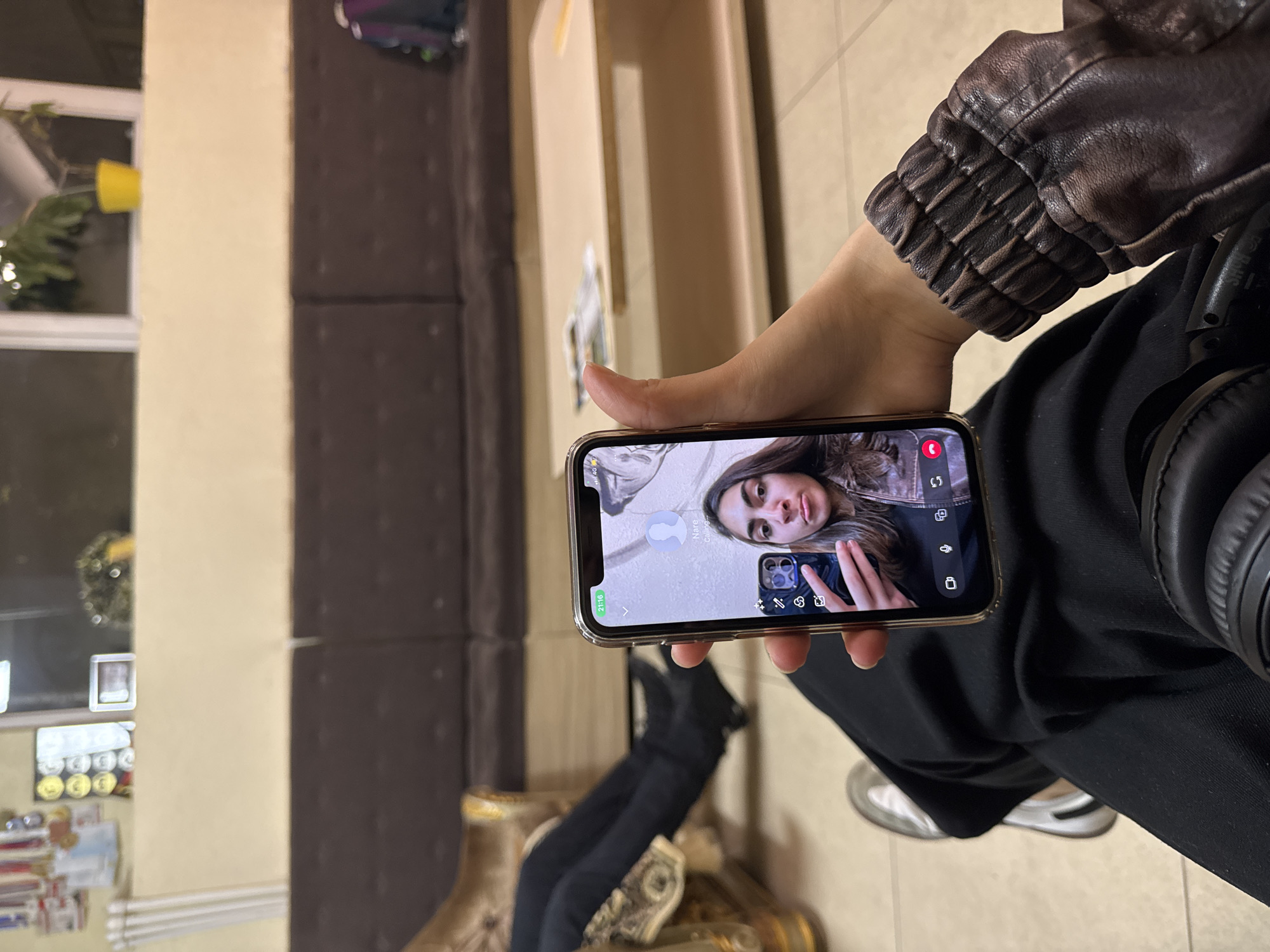 A person sits indoors, holding a gold-colored iPhone in their hand, which displays a video call screen showing a young woman. The person holding the phone is wearing a brown leather jacket and black clothing. A pair of black headphones are placed on their lap.
The background includes a striped wall with alternating bands of beige and brown. A window is visible to the left, with a view of the outdoors and a yellow pot. A desk is visible below the window. A part of the sofa is also visible.
The image was likely taken indoors, possibly in a private residence. I can't determine the location in the city of Yerevan, Armenia, as there are no landmarks or clues that would indicate a specific location.