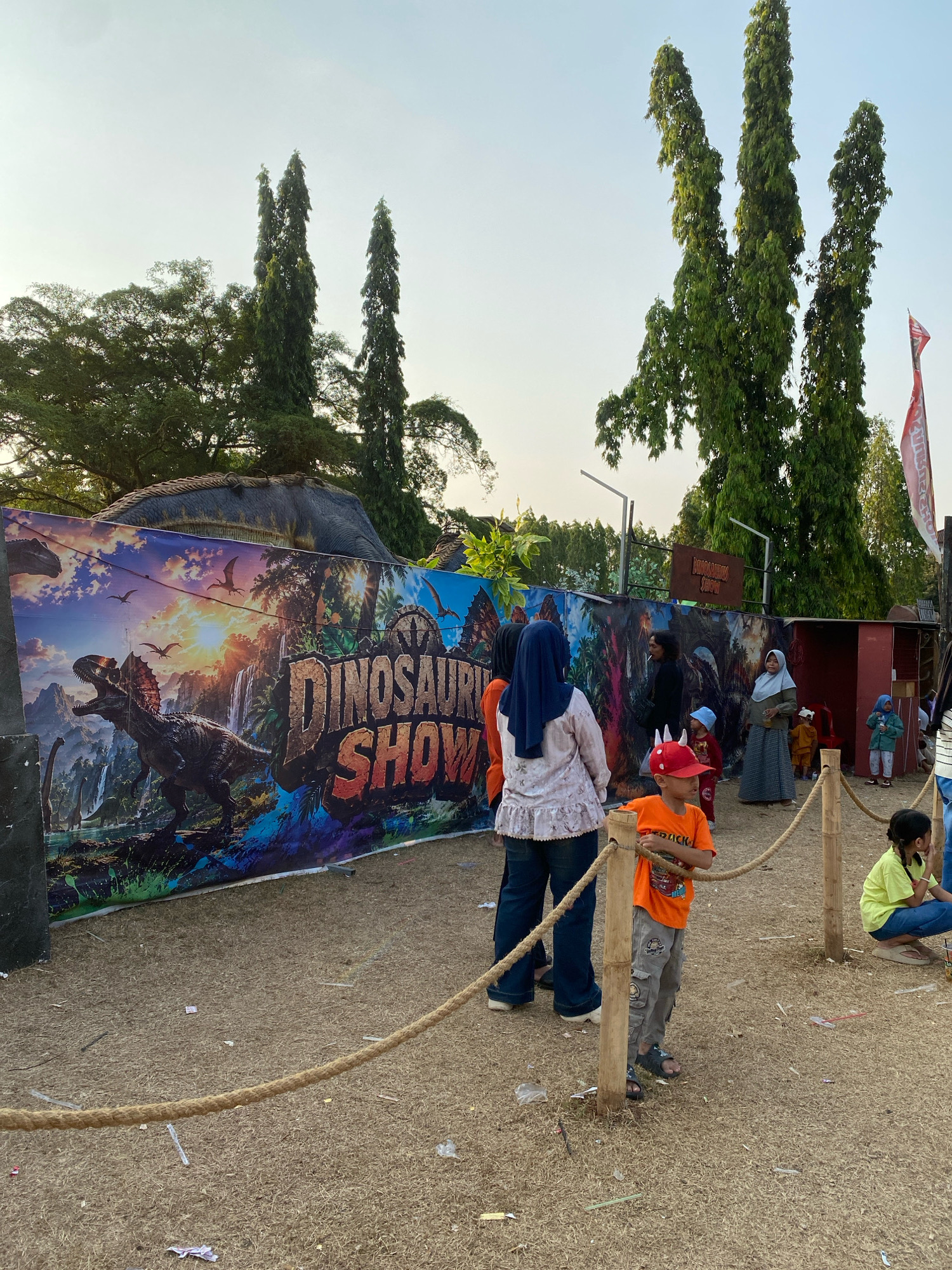 The image captures an outdoor scene at a "Dinosaur Show" event in Nyamok, Indonesia. A large, colorful banner depicting various dinosaurs, lush landscapes, and a waterfall dominates the background. Several tall, slender trees with dense green foliage are visible, along with a clear, pale sky. In the foreground, a group of people, including adults and children, are gathered. One child in an orange shirt and a red hat with horns stands prominently near a bamboo pole, looking towards the right. Another child in a yellow shirt is seated on the ground. A thick, coarse rope stretches across the scene, cordoning off an area. The ground is covered in dry grass and scattered debris. Behind the banner, a large, dark dinosaur structure is partially visible. A red sign with white lettering, possibly indicating the event's name, is mounted on a post. The overall atmosphere suggests a public event or attraction.