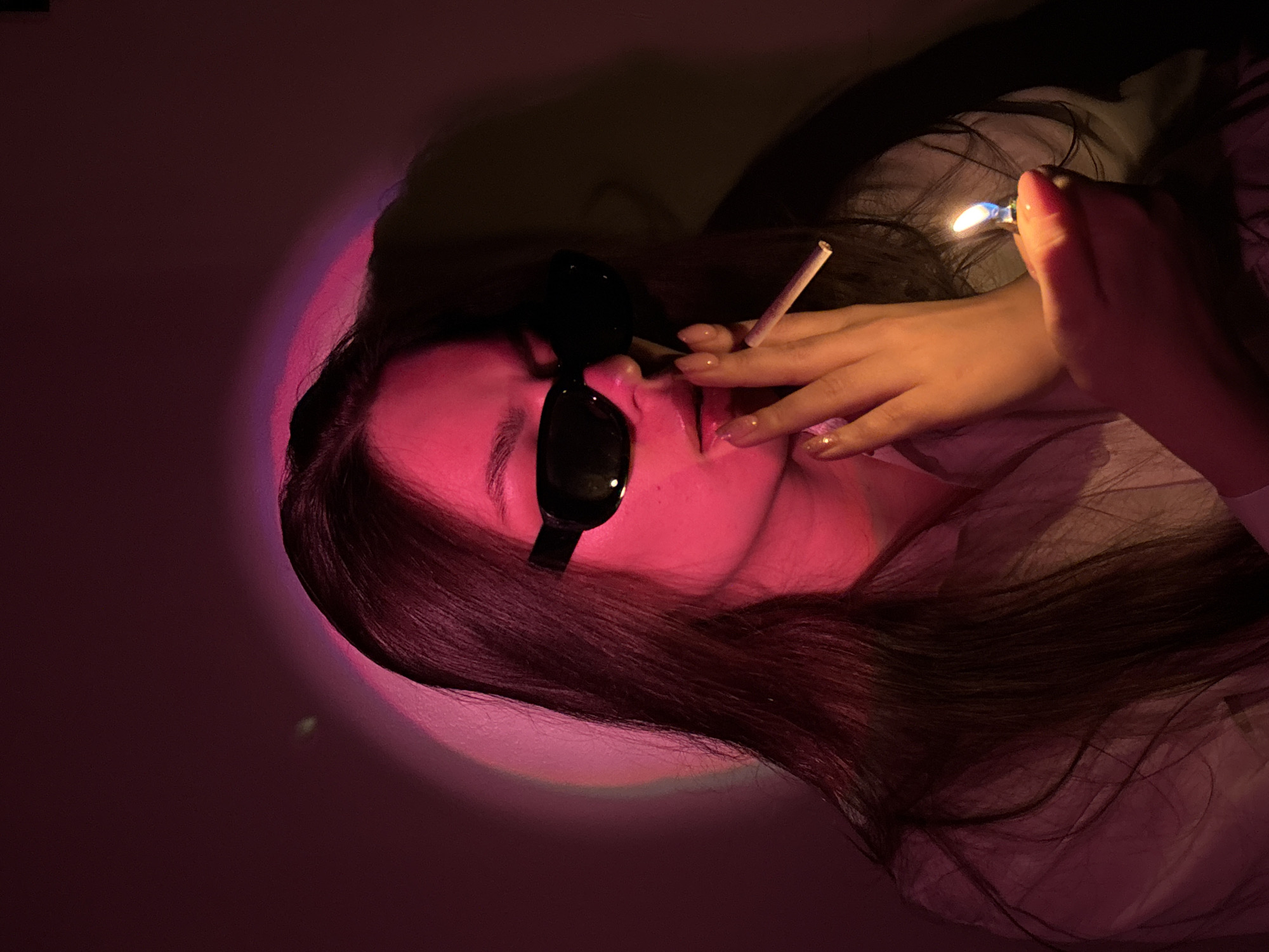 A young woman, with long dark hair, is holding a lit cigarette to her lips while a lighter is held in her right hand. She is wearing sunglasses and a light-colored shirt. The scene is illuminated by a pink light, casting a glow on her face and the surrounding wall. The image does not contain any information that can be used to determine the location within the city of Chaikovsky, Russia, or any other geographical location.