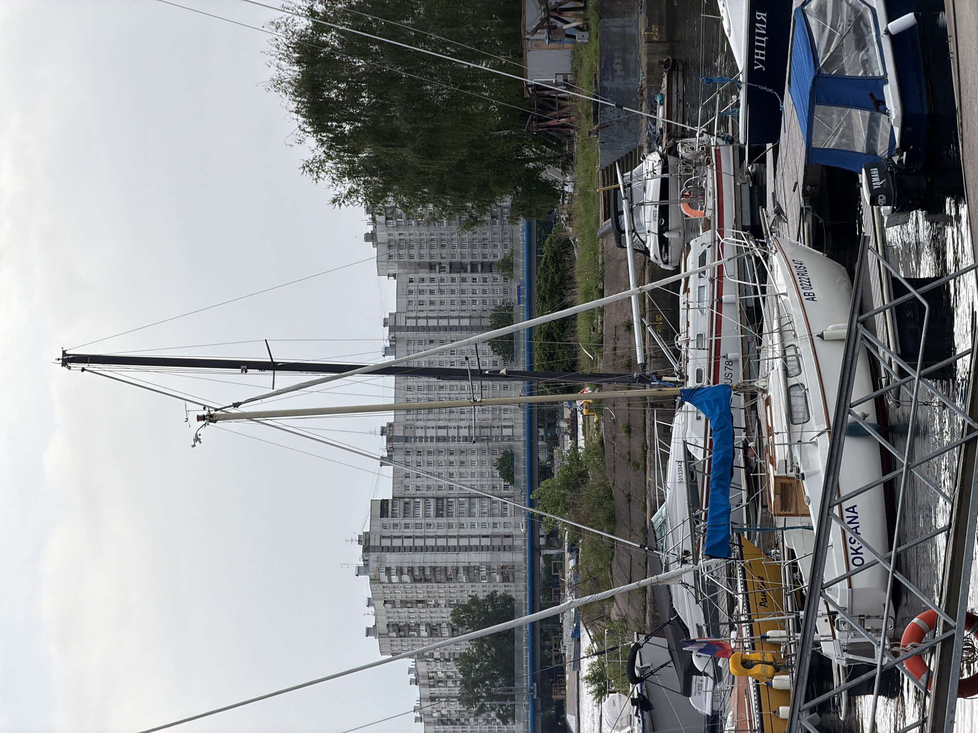 The image depicts a marina in Saint Petersburg, Russia, with several sailboats moored along a dock. The sailboats are of various sizes and designs. Some have their sails furled, while others have portions of their sails unfurled. The names "OKSANA" and "УНЦИЯ" are visible on the sterns of two of the boats. The registration number "AB 0222 RUS 47" is visible on another boat.
In the background are several tall, grey apartment buildings, typical of Soviet-era construction. A tree and some other greenery are also visible between the boats and the buildings. The sky is overcast. A metal railing runs along the edge of the dock. The water is visible between the boats.