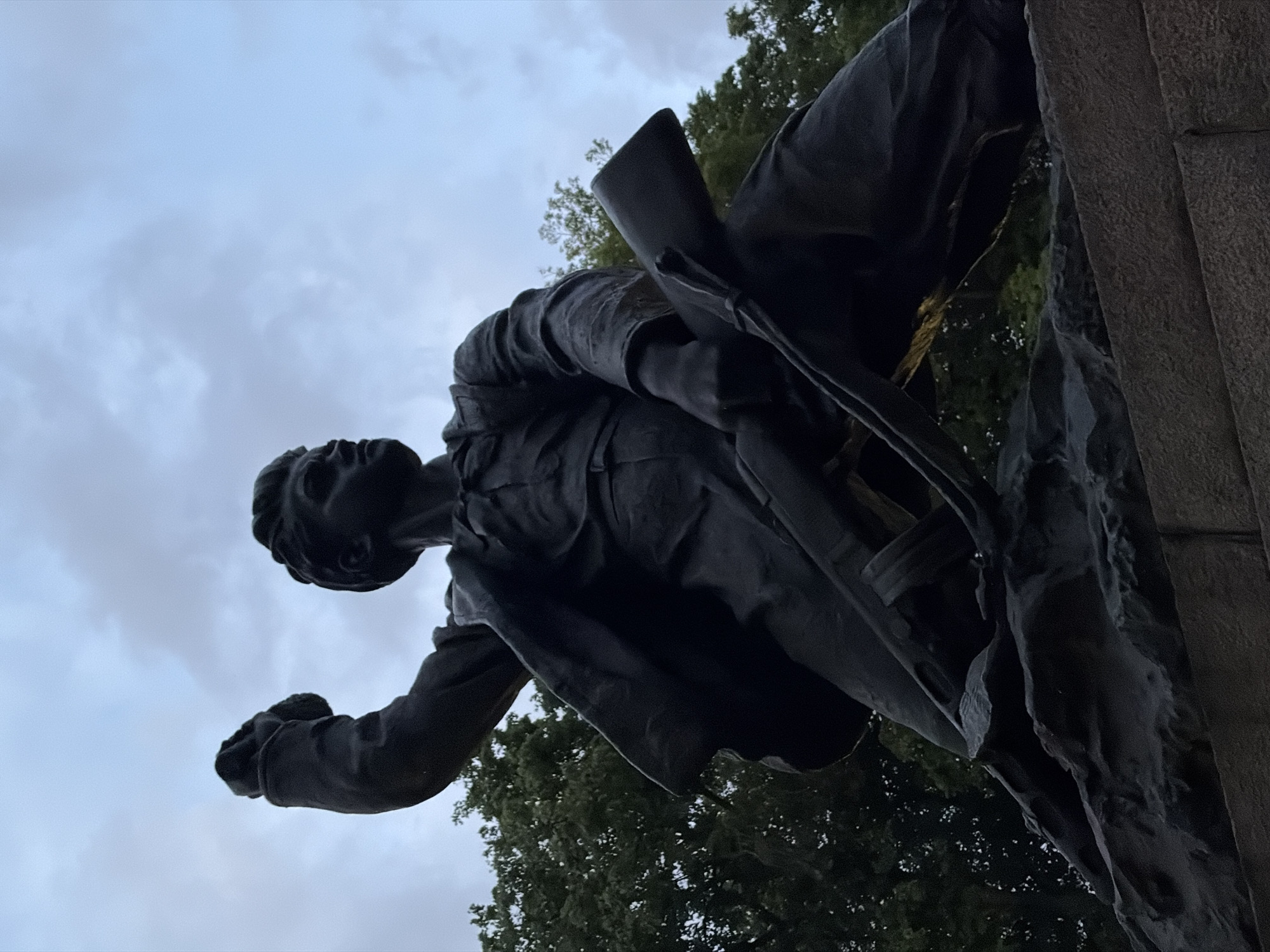 The image depicts a bronze sculpture of a soldier in Minsk, Belarus. The soldier is depicted mid-charge, with one arm extended forward in a fist and the other holding a rifle. He wears a uniform and a determined expression. The sculpture is mounted on a stone pedestal, and the backdrop includes trees and a cloudy sky.