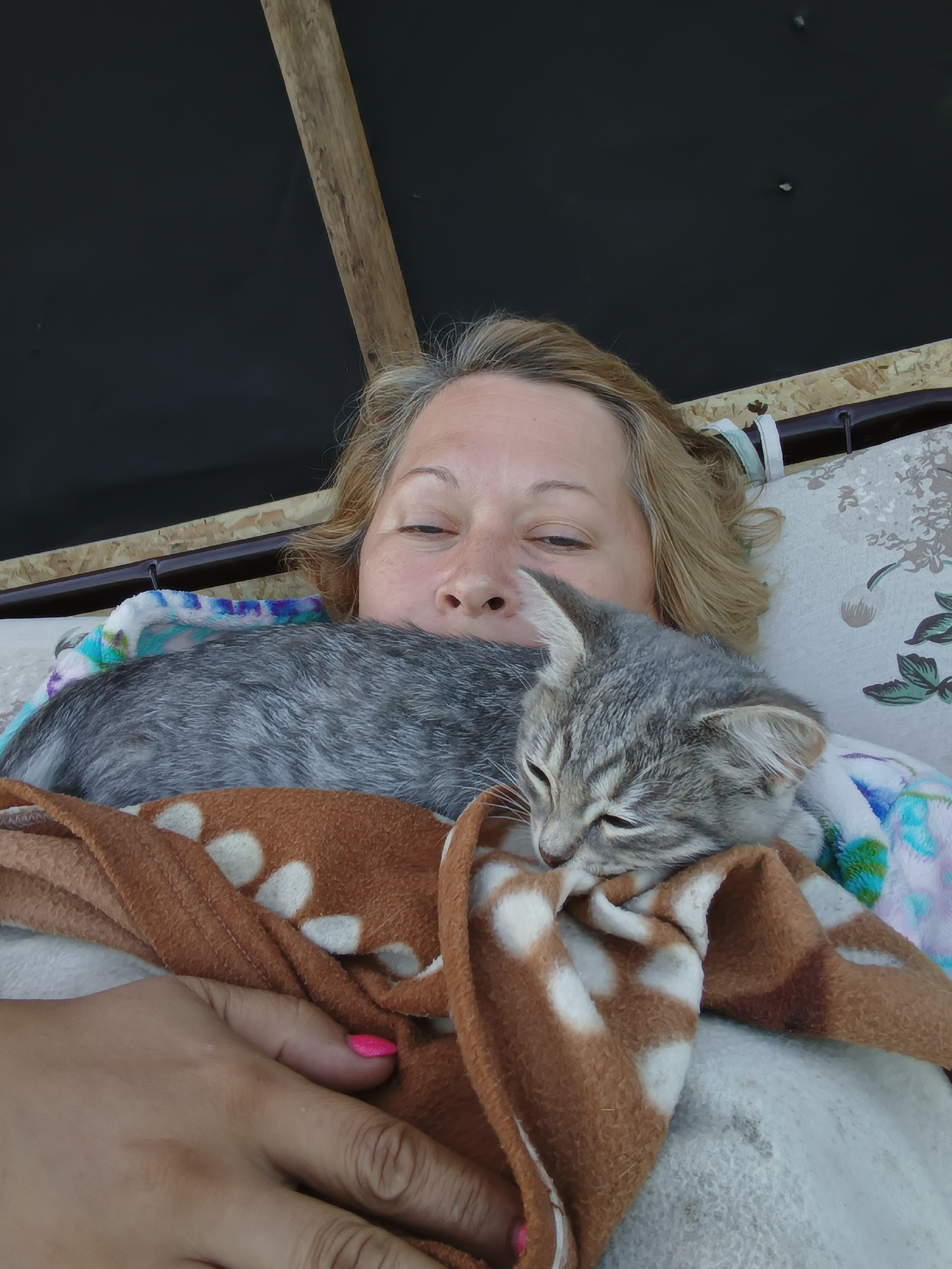 In Ivanovskoe, Russia, a close-up, overhead shot captures a woman and a kitten sharing a blanket outdoors. The woman, with blonde hair, is lying down, her face partially obscured by the kitten. Her eyes are closed or looking down, and she has fair skin with some freckles. A small, grey tabby kitten with striking green eyes is nestled against her, also appearing to be asleep or resting. Both are covered by a brown blanket with white spots and a colorful patterned blanket underneath. The background shows a dark, textured surface with a wooden beam. The scene suggests a moment of rest and connection between the woman and her pet in an outdoor setting.
