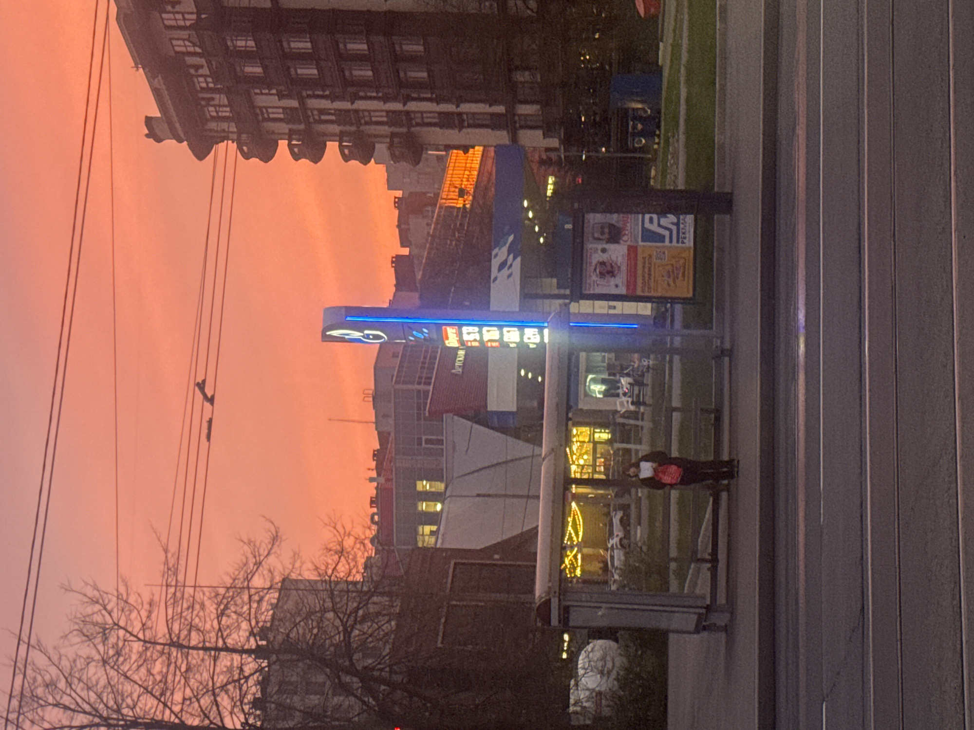 The image depicts a street scene in what appears to be Saint Petersburg, Russia, given the Cyrillic script on the gas station sign. A bus stop, constructed of metal and glass, stands beside a road with parallel vertical lines, possibly marking lanes. A person wearing a light top and dark bottoms stands within the bus stop. Outside, a gas station is visible. The sky is a striking orange, indicating sunset or sunrise. Overhead power lines crisscross the upper left of the frame, and bare trees are silhouetted against the colorful sky. The architecture includes buildings of varying heights and styles, with some appearing older and others more modern.