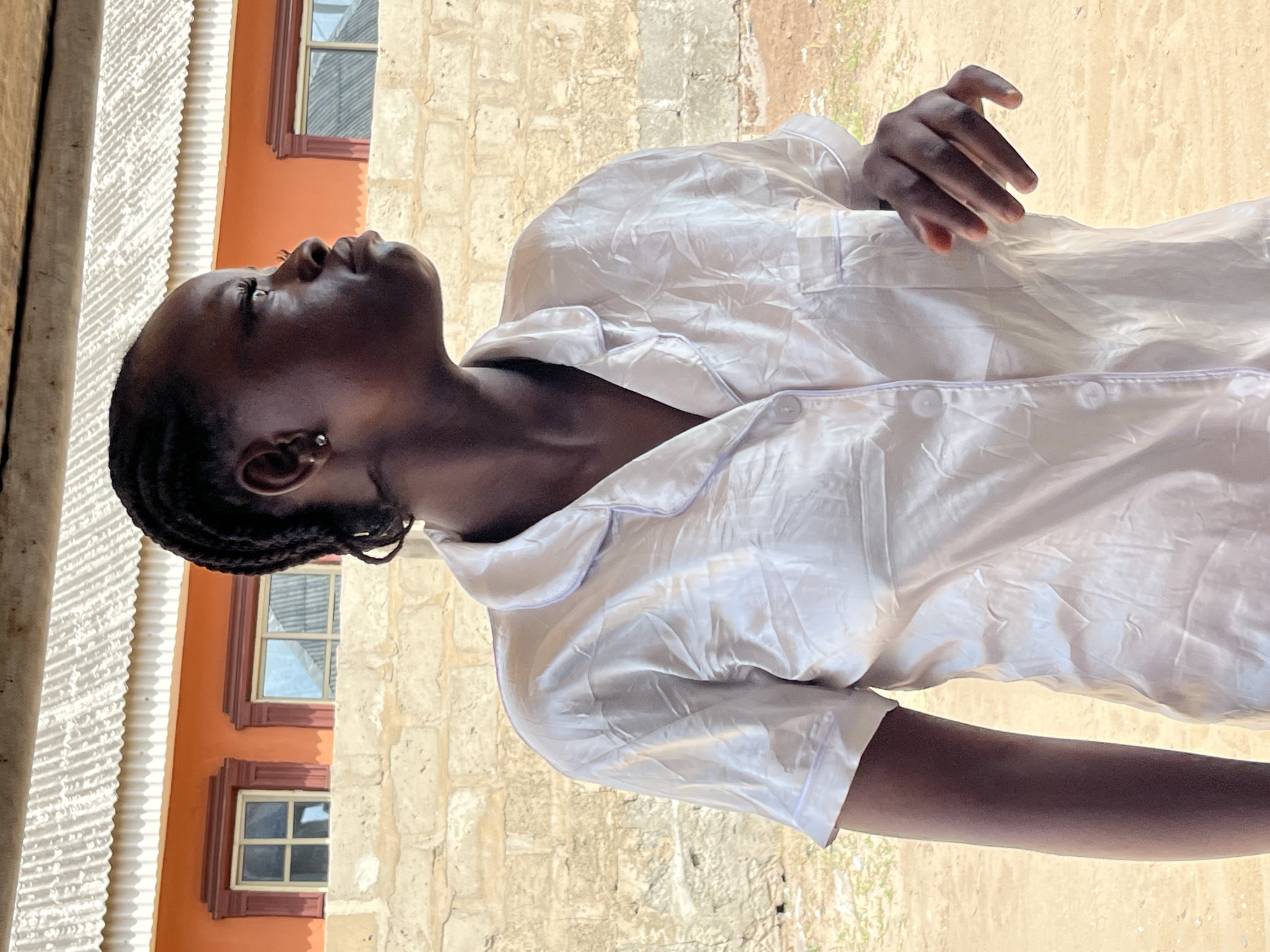A woman with dark skin and braided hair is outdoors in Badagry, Nigeria. She is wearing a white satin pajama set and has a silver earring. She is looking upwards, with a building that has an orange exterior and multiple windows to her left. The building is made of stone. The ground is dirt.
