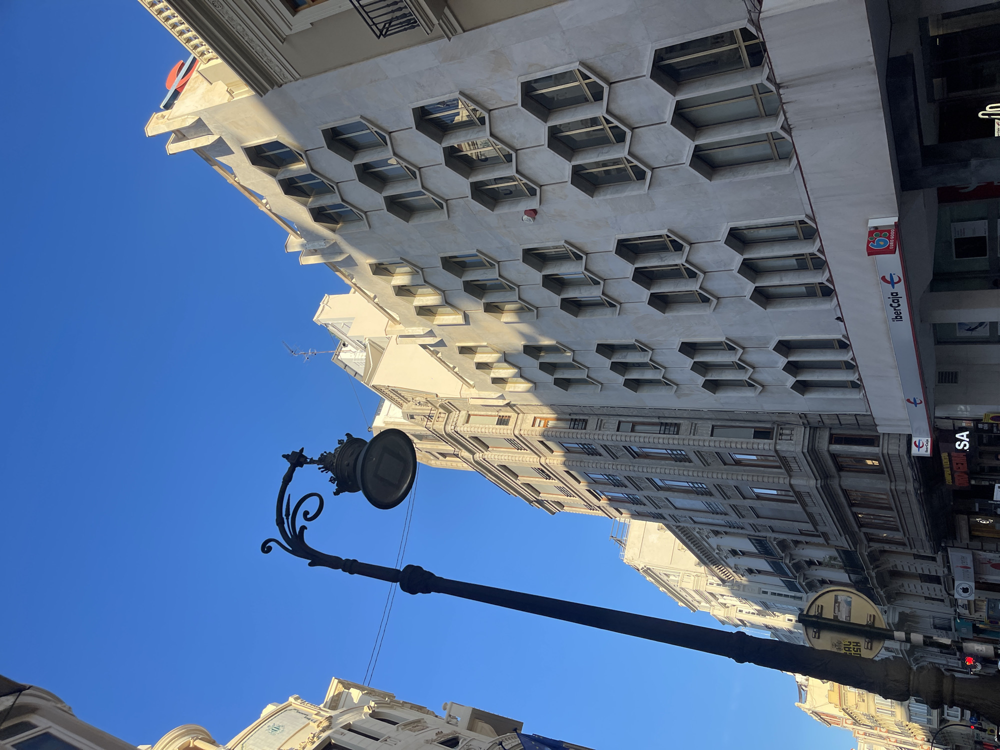The image captures a street scene in Valencia, Spain, looking upward. The sky is a clear, bright blue. Several buildings line the street. One building is notable for its white facade and a honeycomb-like pattern of hexagonal windows. A storefront with the "Ibercaja" logo is visible on one building. A vintage-style black lamppost is prominent in the foreground. The street appears to be somewhat busy, judging by the presence of street signs and other urban elements. The architectural style of the buildings suggests a historic urban setting.