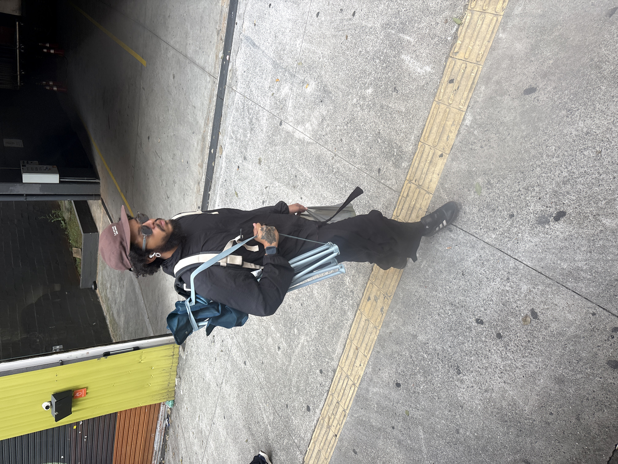 A person wearing a brown baseball cap, sunglasses, and a black outfit walks on a sidewalk next to a yellow and orange corrugated metal building. The person carries a collapsed blue chair strapped to a black backpack. The sidewalk has a tactile paving strip. In the background, a dark tunnel with yellow lines painted on the concrete floor is visible. This scene is likely in a commercial or industrial area of São Paulo, Brazil, given the architecture and urban infrastructure.
