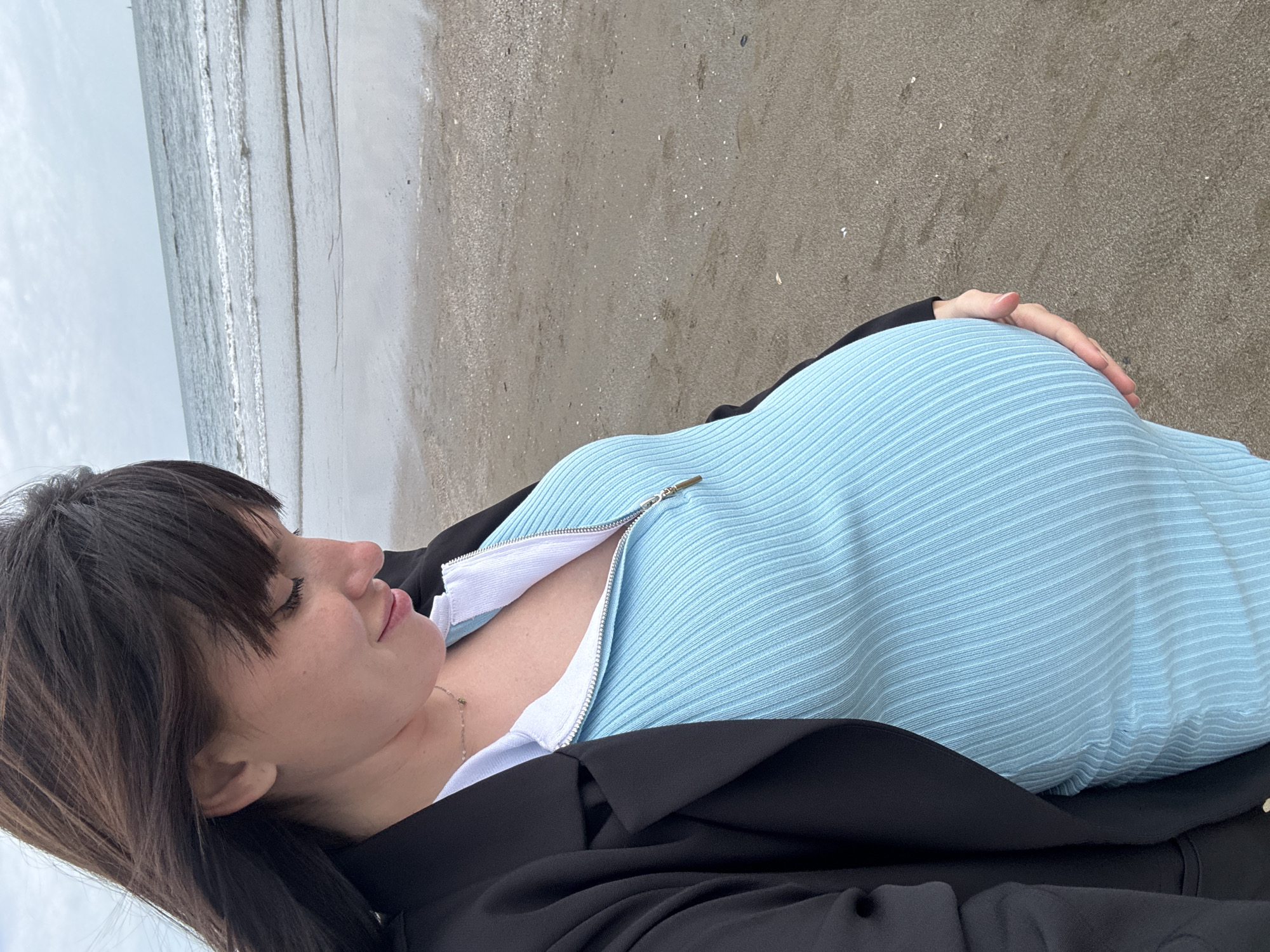 The image captures a serene moment of a woman lying on a sandy beach in Nettuno, Italy, under an overcast sky. The photo is taken from an overhead perspective, with the woman positioned horizontally across the frame.
The main subject is a woman with dark hair and bangs, seen in profile with her eyes closed or gazing downwards, exhibiting a peaceful and relaxed expression with a subtle smile. She is dressed in a light blue, ribbed top or dress that features a white zipper, partially unzipped, revealing a delicate gold-colored chain necklace. A black jacket or blazer is draped over her shoulders and torso. Her rounded midsection suggests pregnancy, and one of the cropped images shows her hand gently resting on her belly, adding to the tender and calm atmosphere.
The setting is a tranquil beach environment. The foreground consists of damp, grey-brown sand, marked by ripples from receding waves and scattered small detritus. In the background, the calm, light blue-grey sea meets the shore, with gentle waves creating white lines along the waterline. The sky above is a uniform light grey, indicating an overcast day.
The overall impression is one of quiet contemplation and natural beauty. There is no visible text in the image.