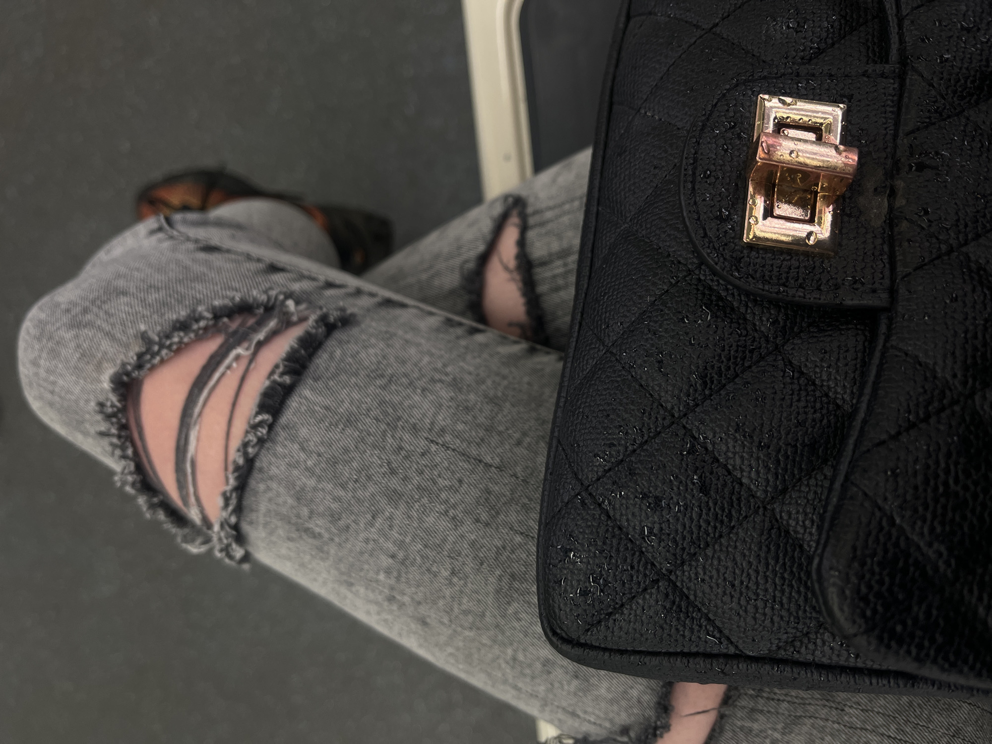 The scene captures a close-up, downward-angled view of a person's lower body and a handbag inside what is consistent with a public transport vehicle, such as the Sofia Metro or a bus, in Sofia, Bulgaria. The individual is seated with legs crossed, wearing faded grey denim jeans that feature prominent horizontal rips across both knees, exposing the skin beneath. A black, textured, quilted handbag occupies the foreground, presenting a gold-colored rectangular turn-lock clasp. Numerous small water droplets are visible across the handbag's surface, including its textured material and the metal clasp. A blurred, dark-toned shoe, likely brown, is partially discernible on the individual's right foot. The background consists of a dark grey floor or surface, with a light-colored horizontal structural element visible on the right, indicative of an interior panel or seating component of the vehicle.