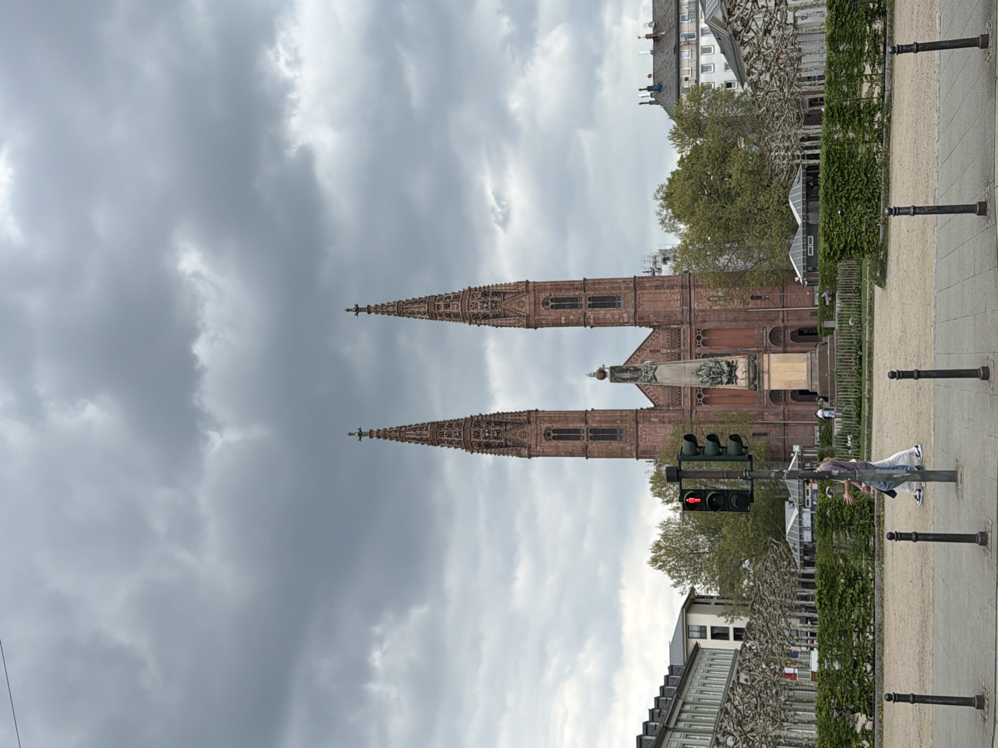 The image shows a sideways view of the Market Church (Marktkirche) in Wiesbaden, Germany. The sky is overcast with dark gray clouds. The church features two prominent spires made of red brick. A statue is visible in front of the church. A traffic light shows a red signal. On the right side of the frame, a person in casual clothing is leaning against a pole and appears to be stretching. A green hedge, trees and other buildings are also visible. The ground appears to be a combination of pavement and gravel. Black metal posts line the walkway.