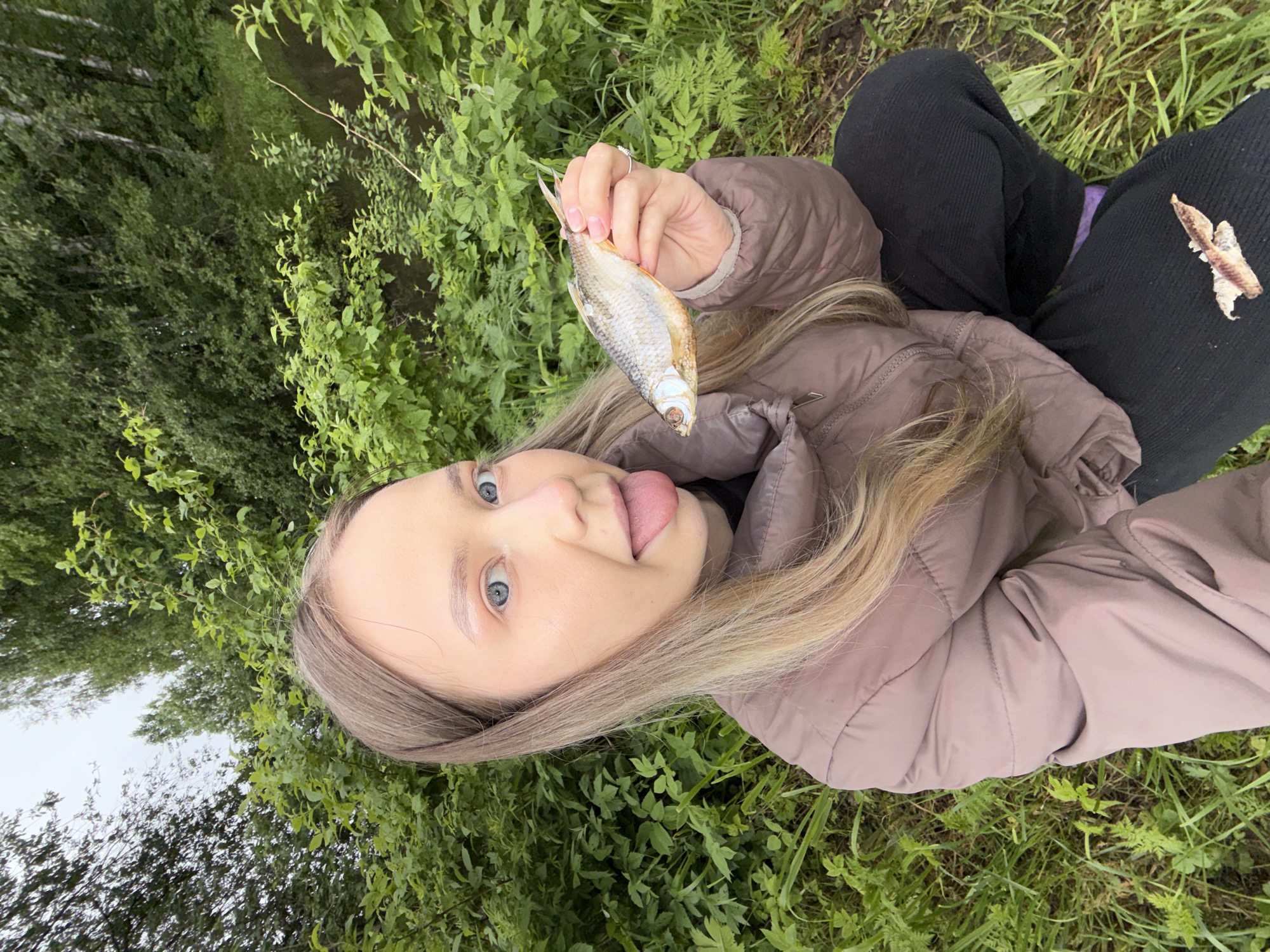 A young woman is outdoors in a grassy area, possibly near a body of water implied by the surrounding foliage. She is holding up a small, dried fish in her right hand. She is wearing a light brown puffer jacket and dark pants. She has long blonde hair and blue eyes. She is sticking her tongue out. A fragment of what appears to be another fish lies on her pants. The location is Zhigalki, Russia.