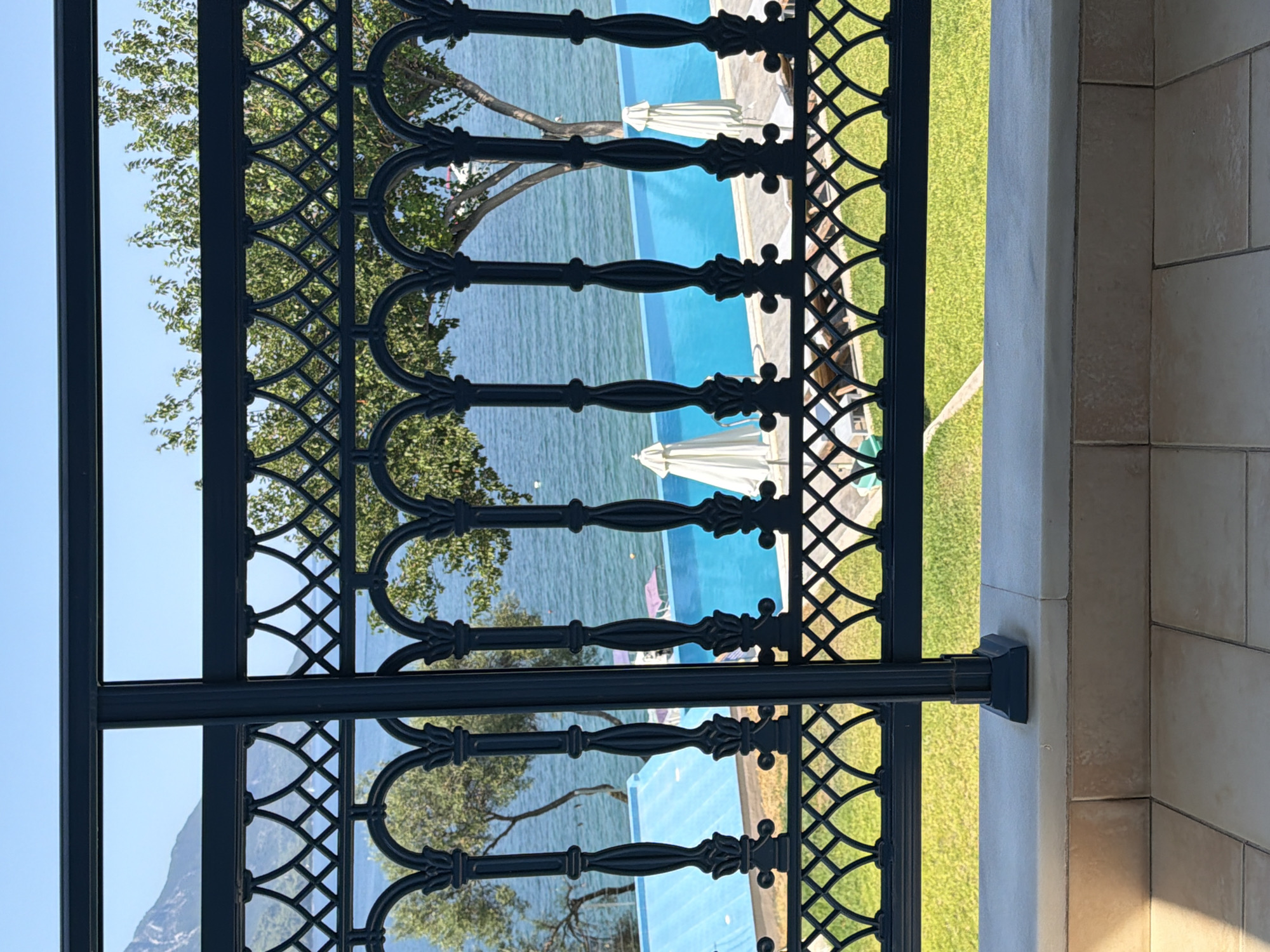 This is a photograph of a view from a balcony overlooking the sea in the Municipal Unit of Alyzia, Greece. A black wrought-iron railing with an ornate design runs across the foreground, framing the scene. Beyond the railing, a bright blue swimming pool is visible, with the calm sea stretching to the horizon. Trees with green foliage are scattered along the coastline. The sky is clear and blue. The balcony itself has a light-colored stone ledge and tiled flooring. There are lounge chairs and umbrellas around the pool, suggesting a resort or hotel setting. The overall impression is one of a serene and luxurious vacation spot.