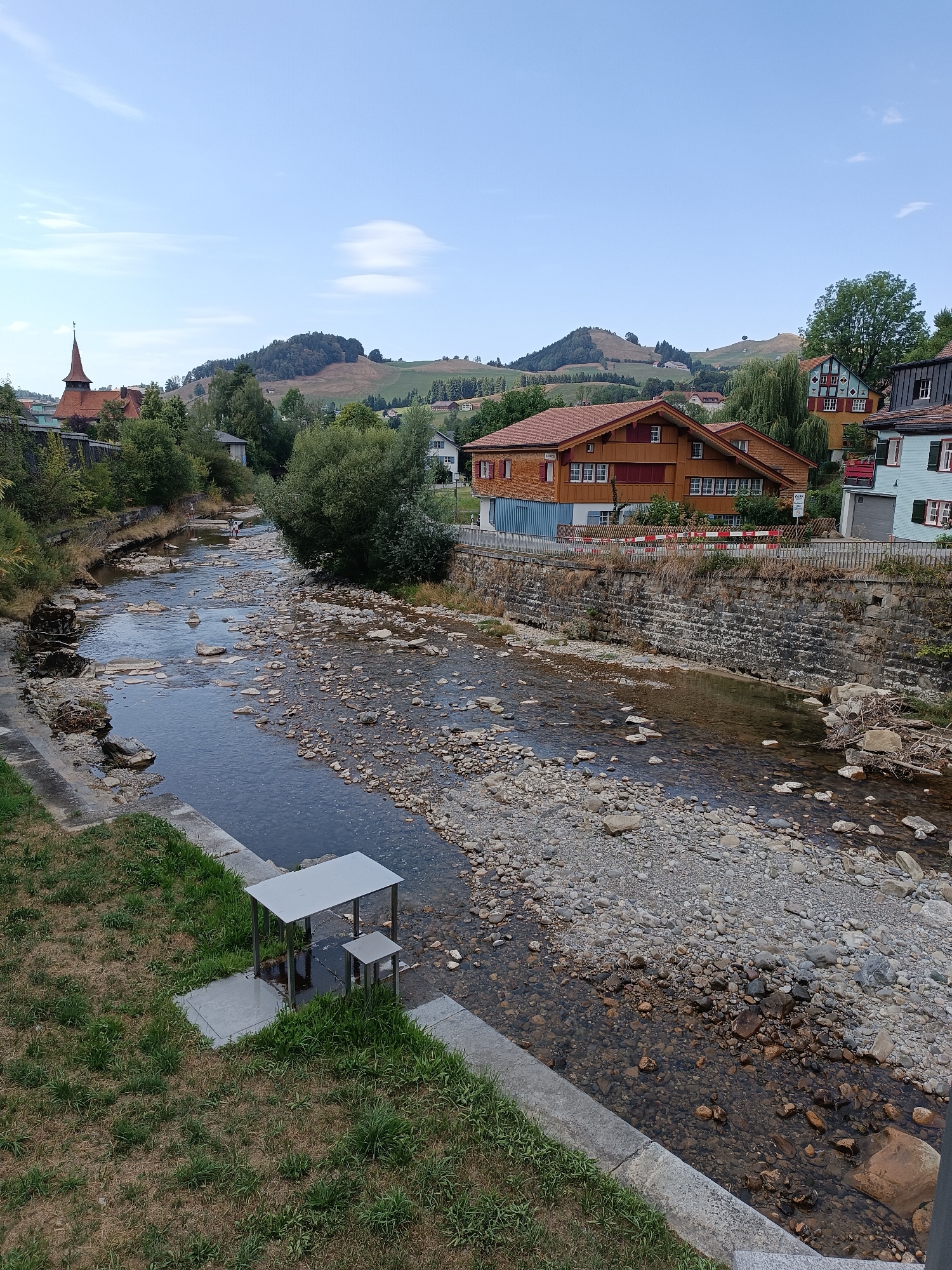 The image captures a view of the river running through the town of Appenzell, Switzerland. The riverbed is rocky and shallow, with clear water flowing over the stones. On the left bank, there is a grassy area with a modern metal table and bench set up, situated on a concrete base. Further back, a stone wall lines the river, and beyond that, buildings and trees are visible.
On the right bank, several houses are depicted. One prominent building is a two-story house with a distinctive orange-red wooden exterior and a steep, tiled roof. It has numerous windows with white frames and red shutters. Next to it, a lighter colored house with blue and yellow accents can be seen. The houses have traditional Swiss architectural features, including wooden balconies and gabled roofs.
In the background, rolling green hills covered in trees and some patches of grass rise up, indicating the rural setting surrounding the town. The sky is clear and blue with a few wispy clouds. The overall scene suggests a peaceful, picturesque town nestled in a valley with a river flowing through it. A steeple of a church can be seen on the far left side of the image, adding to the town's character. In the river itself, a few people can be seen wading or playing in the water, indicating the river is used for recreation.